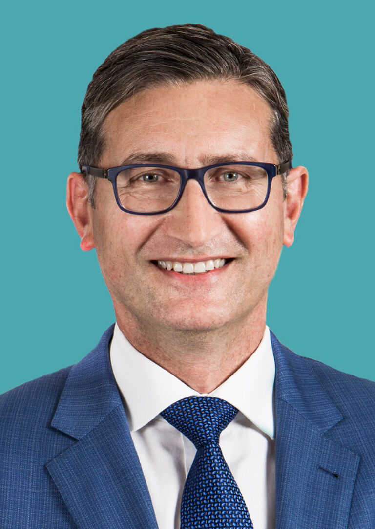 A smiling businessman with dark hair, wearing glasses, a navy suit, and a blue tie against a teal background.