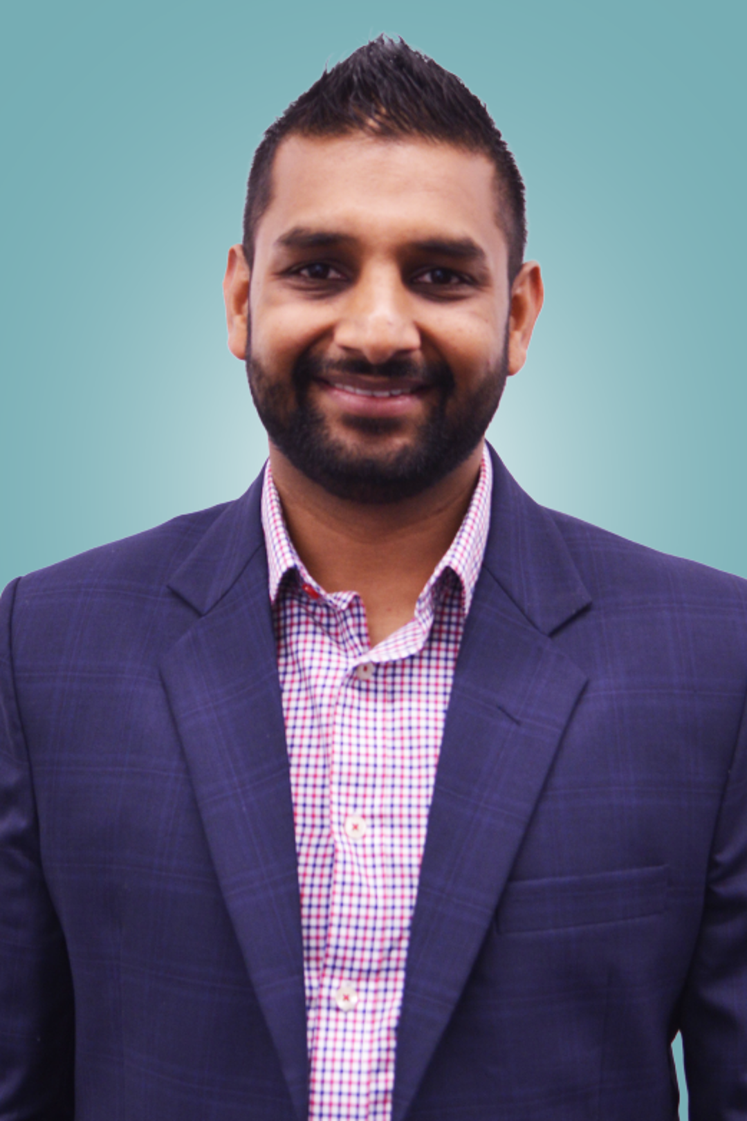 A well-groomed man with a beard smiles confidently in a professional setting, wearing a navy blazer over a checkered shirt.