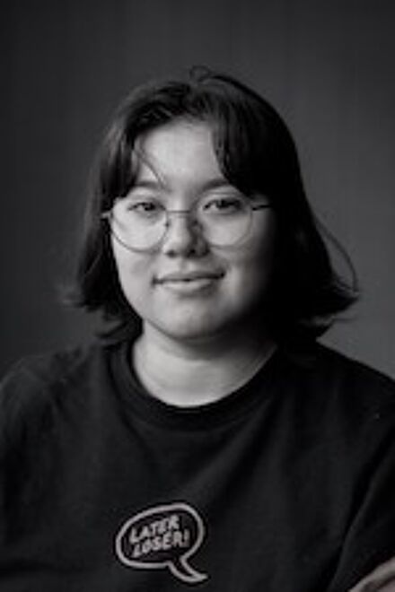 A black-and-white photograph capturing a cheerful young woman with mid-length hair wearing round glasses and a t-shirt with a slogan.