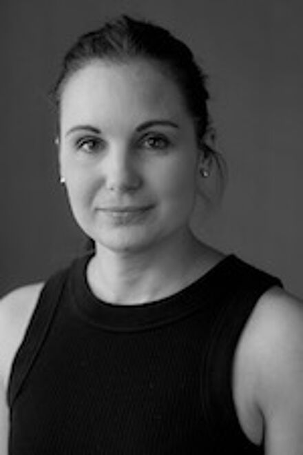 A black-and-white headshot of a woman with her hair tied back, wearing a sleeveless top and modest earrings, exuding confidence and professionalism.