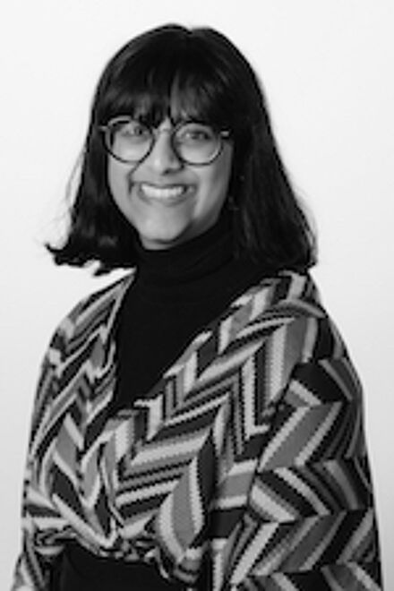 A cheerful woman with glasses, wearing a patterned shawl, poses for a black and white portrait.