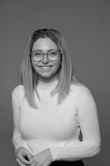 A grayscale image of a cheerful woman with long straight hair wearing glasses and a white turtleneck, standing confidently.