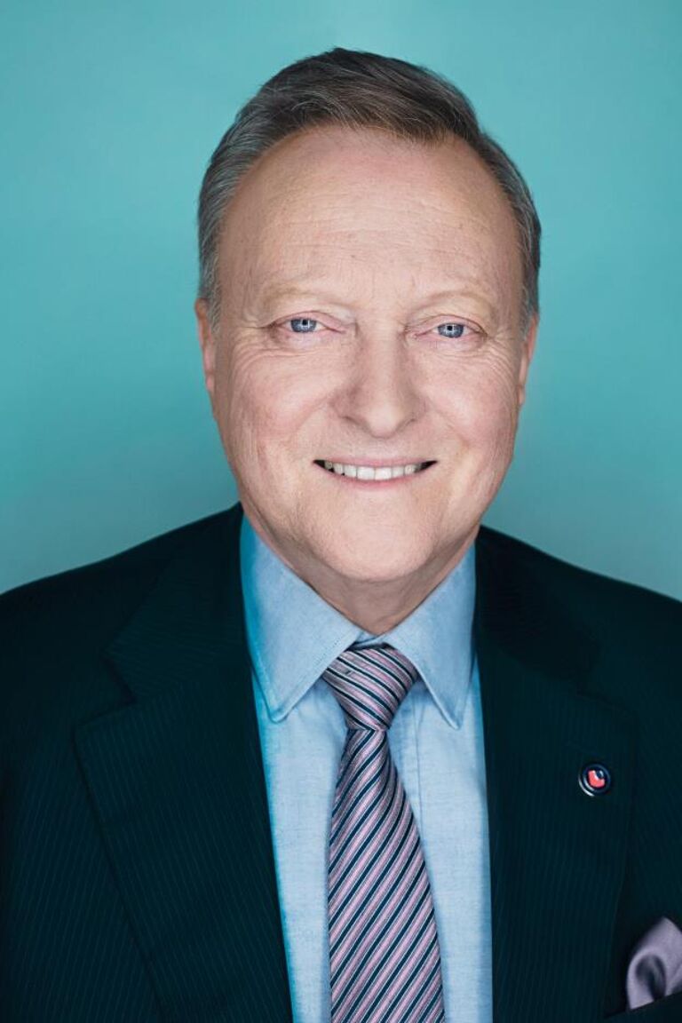 Portrait of an elderly man with a warm smile, wearing a dark suit, striped tie, and a light blue shirt against a teal background.