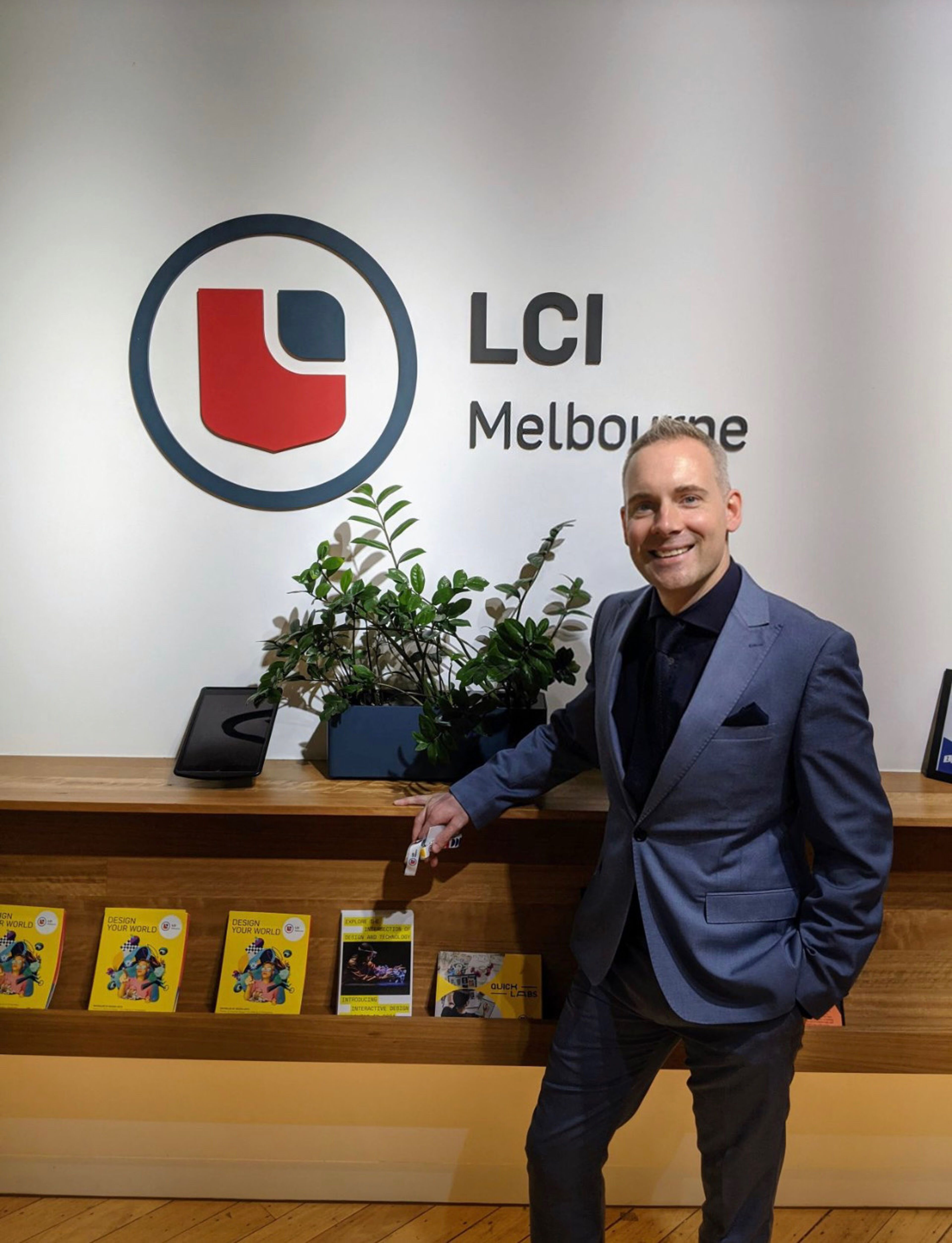 Un individu souriant se tient fièrement devant le logo de LCI Melbourne, symbolisant l'accueil chaleureux de l'institution.