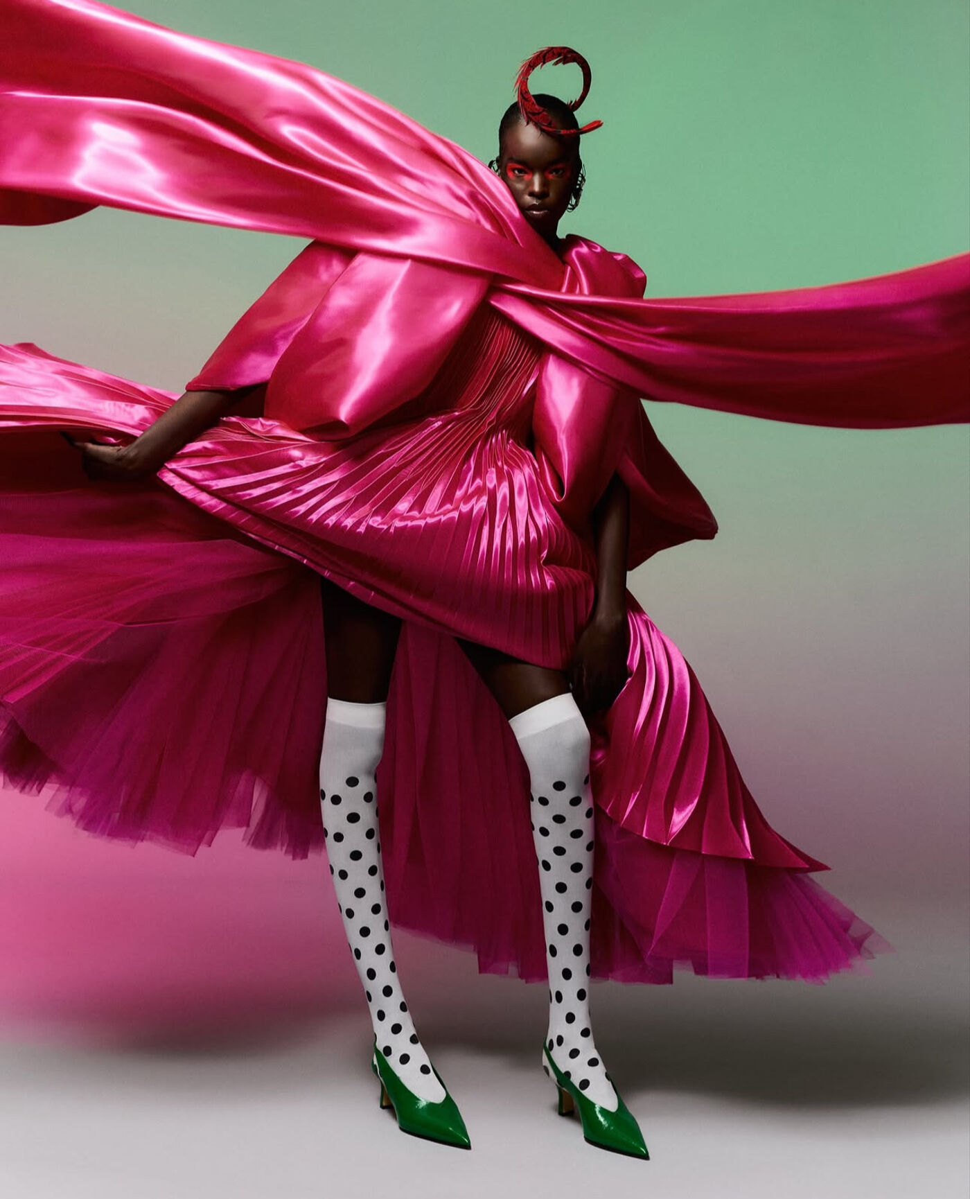 A person with dark skin is wearing a flamboyant, bright pink dress with a large, flowing cape. They are also wearing white knee-high socks with black polka dots and green pointed shoes.