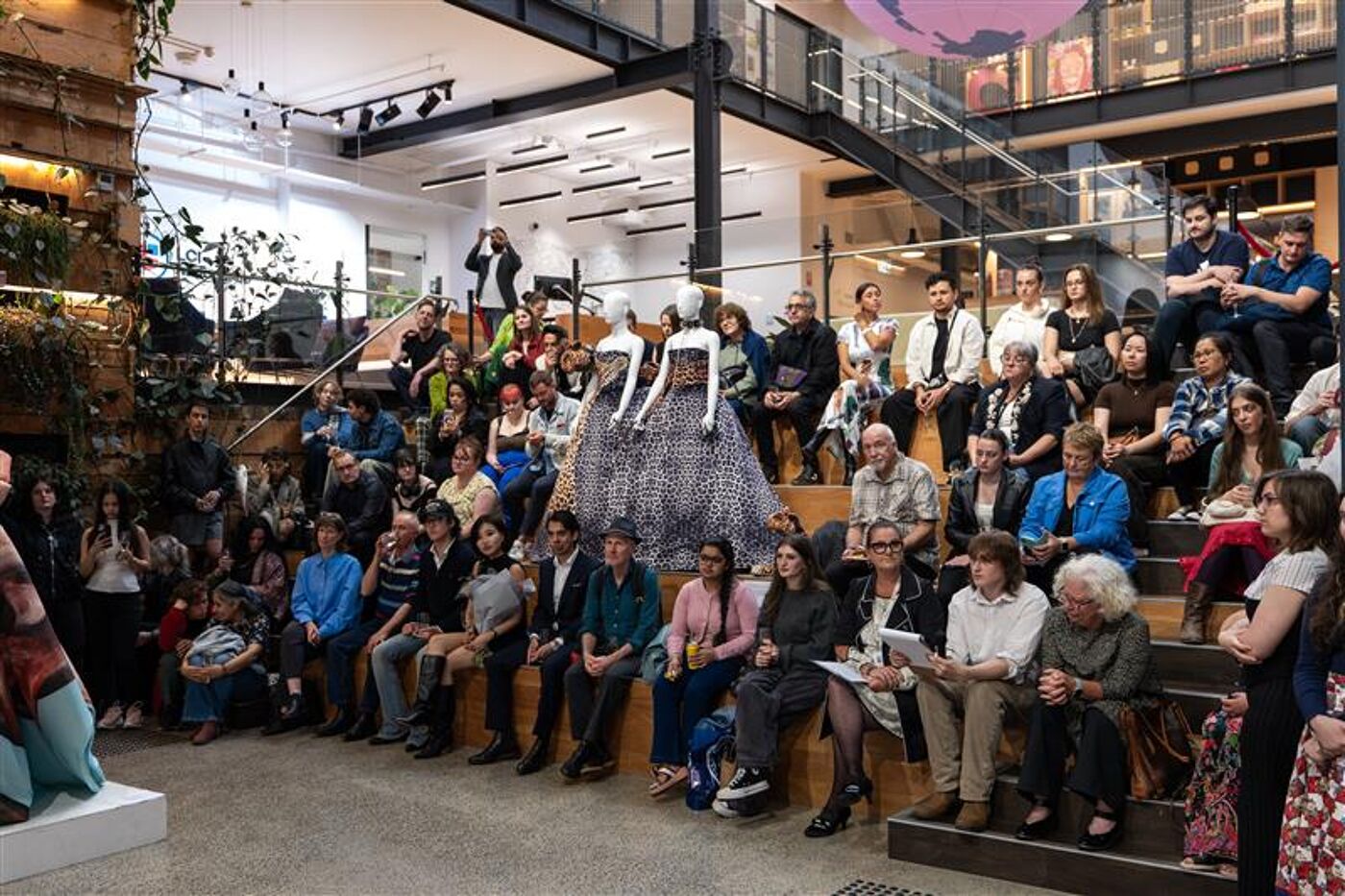 A crowd sits in a modern atrium, observing a performance with figures dressed in white and grey outfits before them.