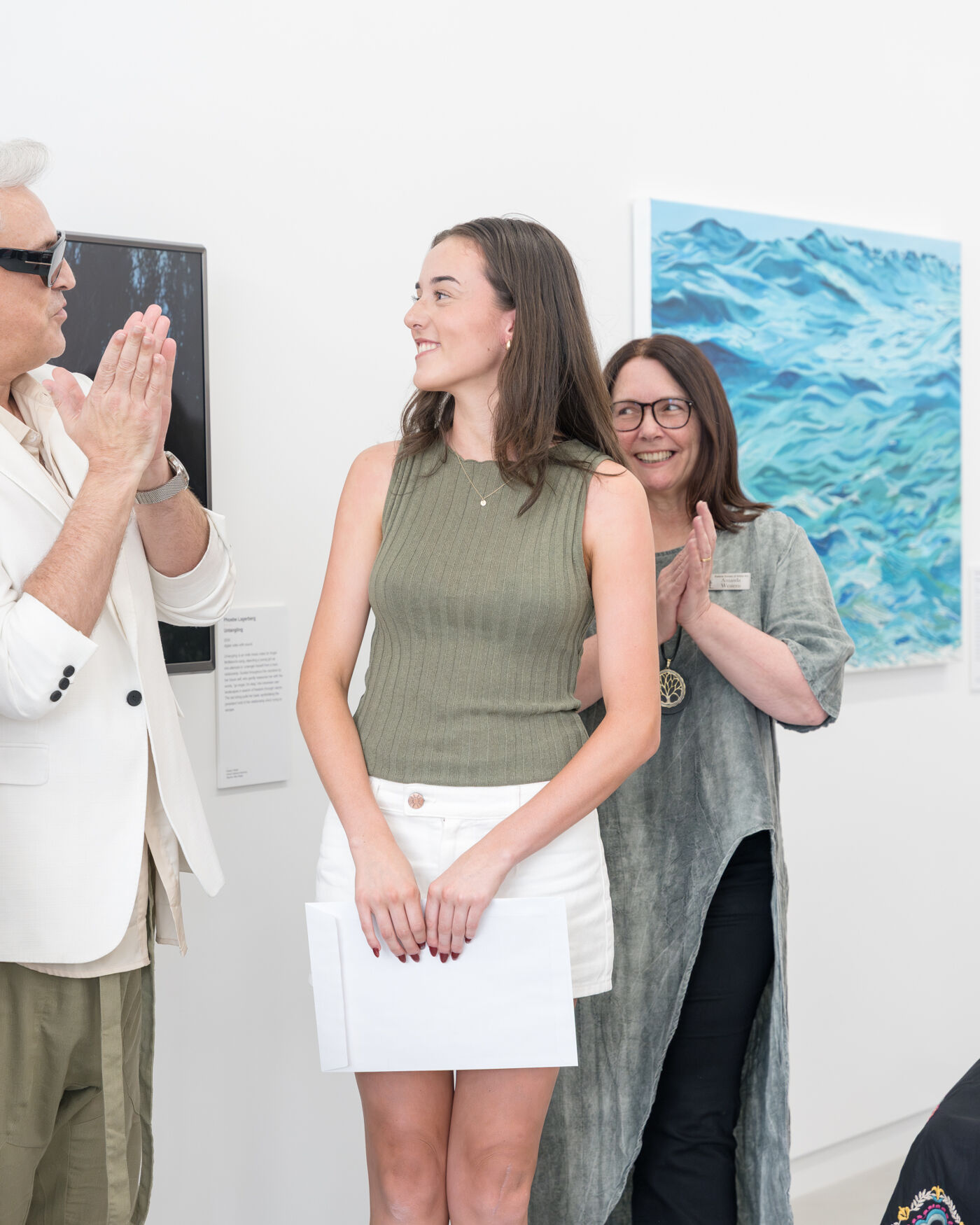 Three people are in an art gallery, two women and a man wearing sunglasses, clapping with their hands together.
