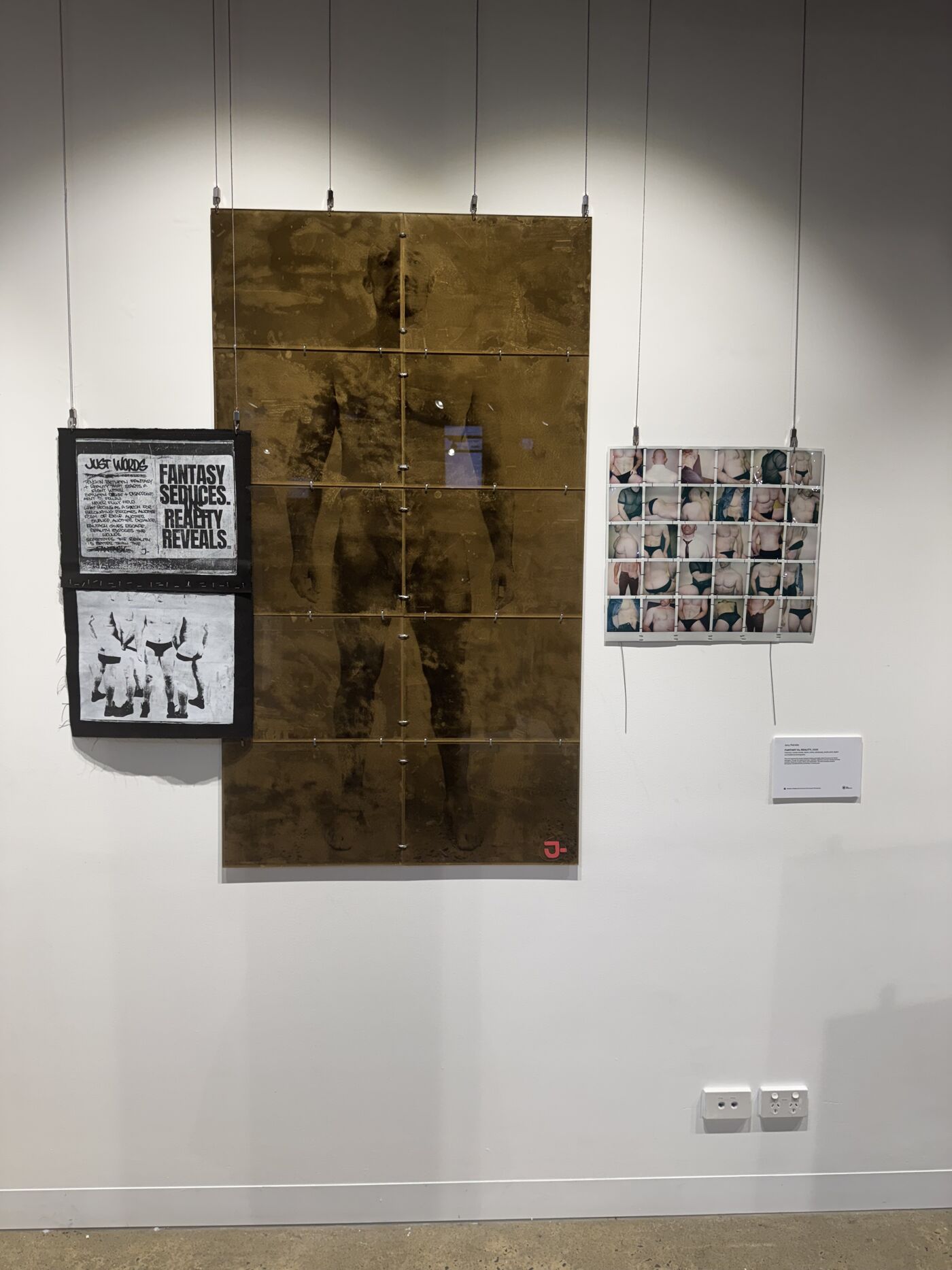 This image displays an art exhibition featuring three pieces hanging on a white wall. The central piece is large and golden, showing the silhouette of a human figure. To the left is a framed artwork with black and white text and images. To the right is a small collage arranged in a grid.