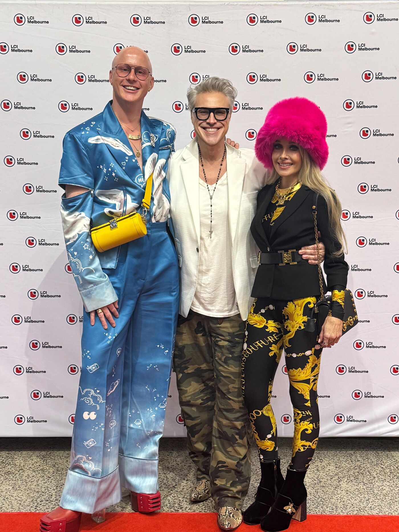 Three individuals pose in front of a backdrop featuring the 'Life Melbourne' logo. The person on the left wears a silky blue outfit with cut-out sleeves, the middle person sports a white jacket and camouflage pants, and the person on the right is dressed in a black jacket, patterned pants, and a bright pink hat.
