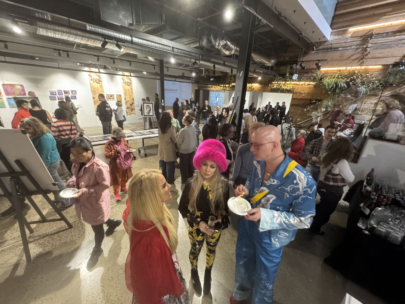 A crowd of people attends an art exhibition, with various artworks displayed on the walls and easels set up for live art creation.