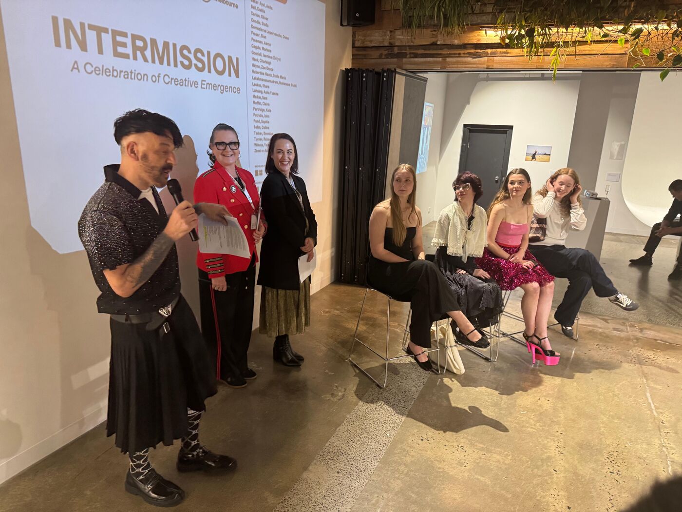 A man in a kilt and a group of young women are at an event with the word "INTERMISSION" on the screen.