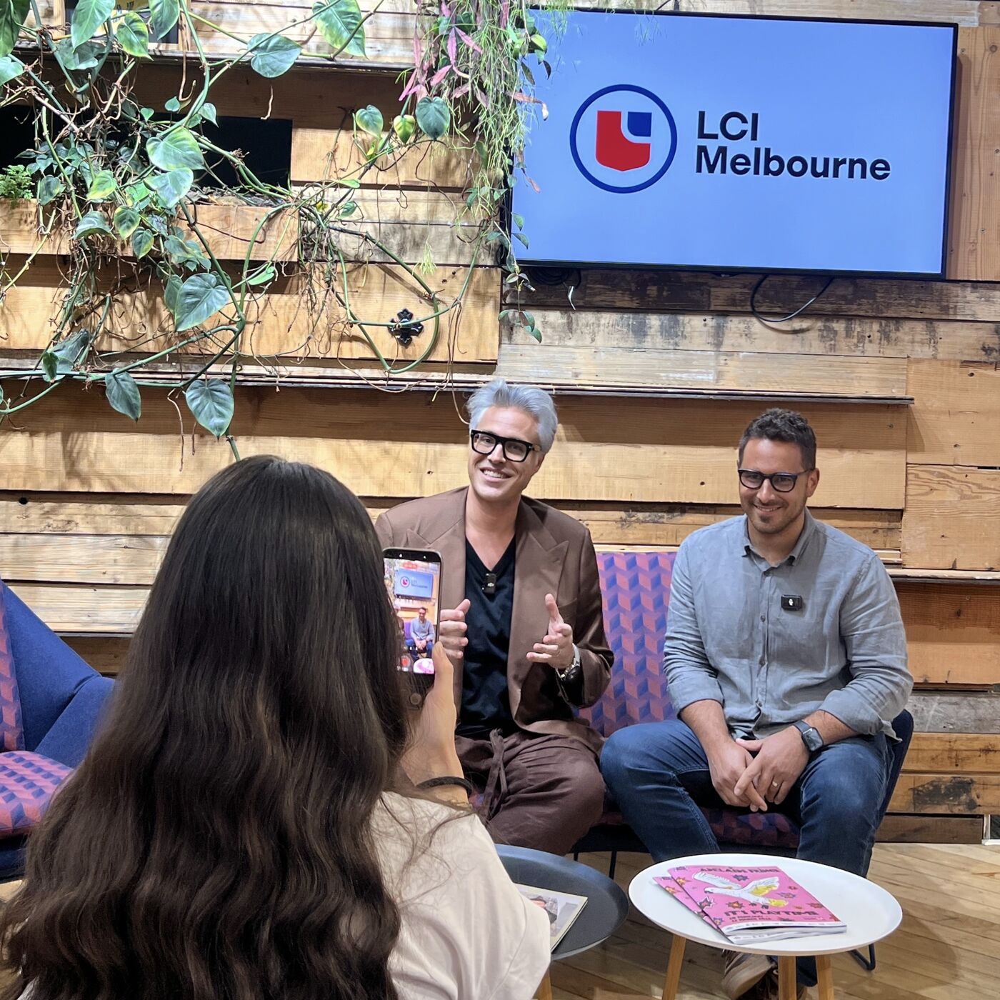 Two people are seated on a couch while a third person films with a phone. In the background, a screen displays "LCI Melbourne".