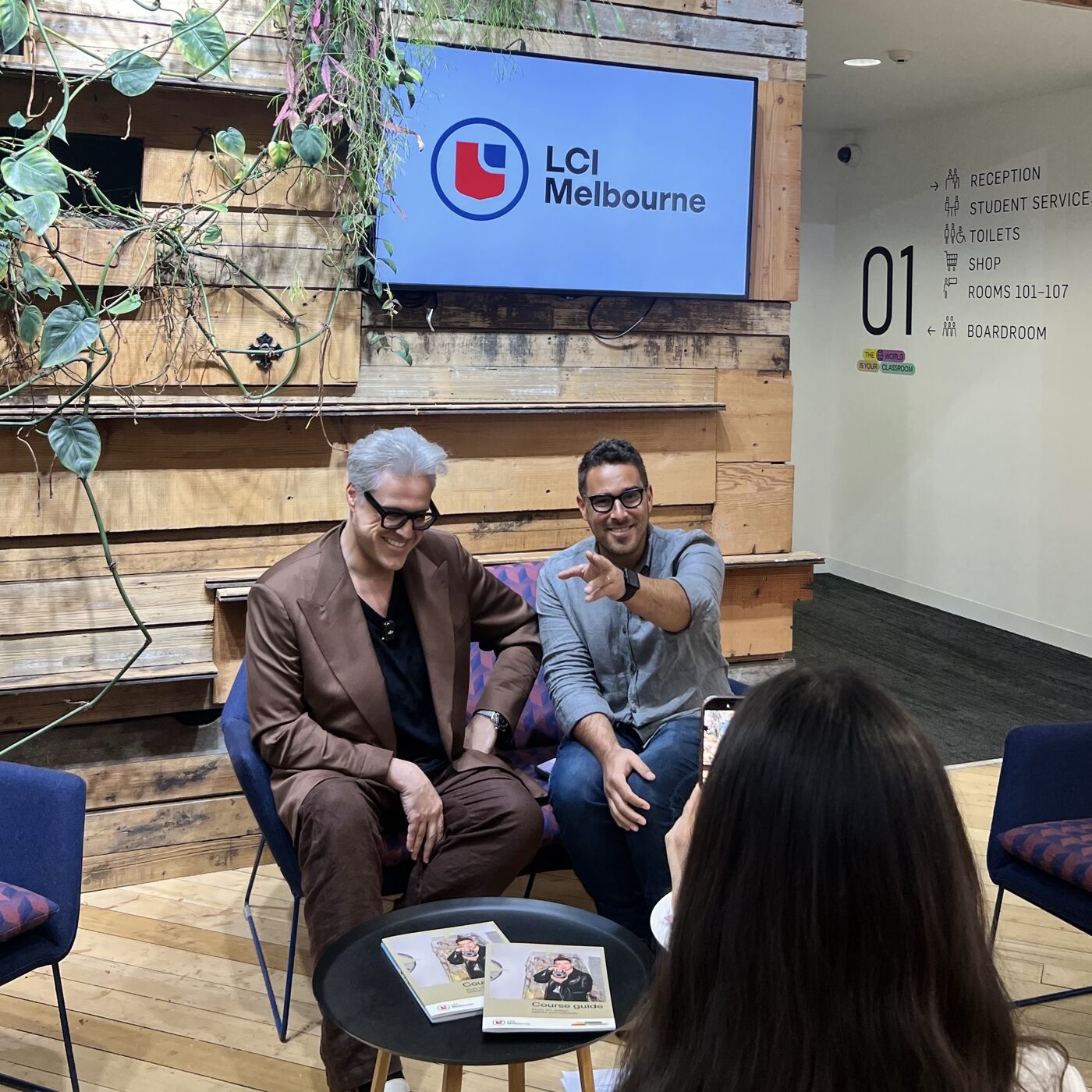 Two people are seated in front of a table with books, with an LCI Melbourne logo in the background. One man is pointing towards the camera.