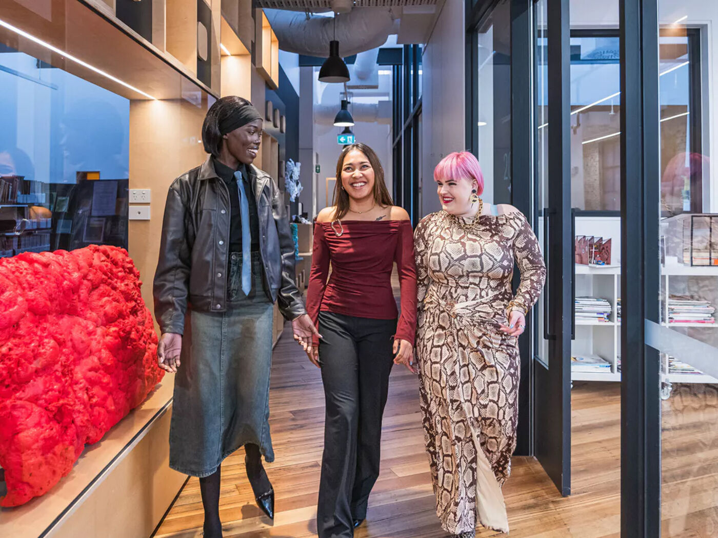 Three diverse women walk together down a modern hallway, smiling and interacting positively, creating a professional yet welcoming atmosphere.