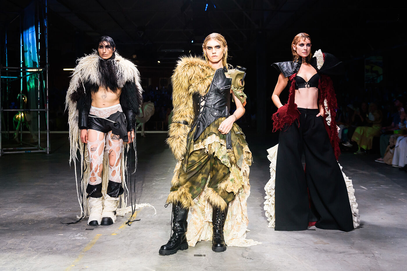 Three models showcase avant-garde, post-apocalyptic-inspired outfits on a runway, featuring dramatic textures, faux fur, and unique layering.