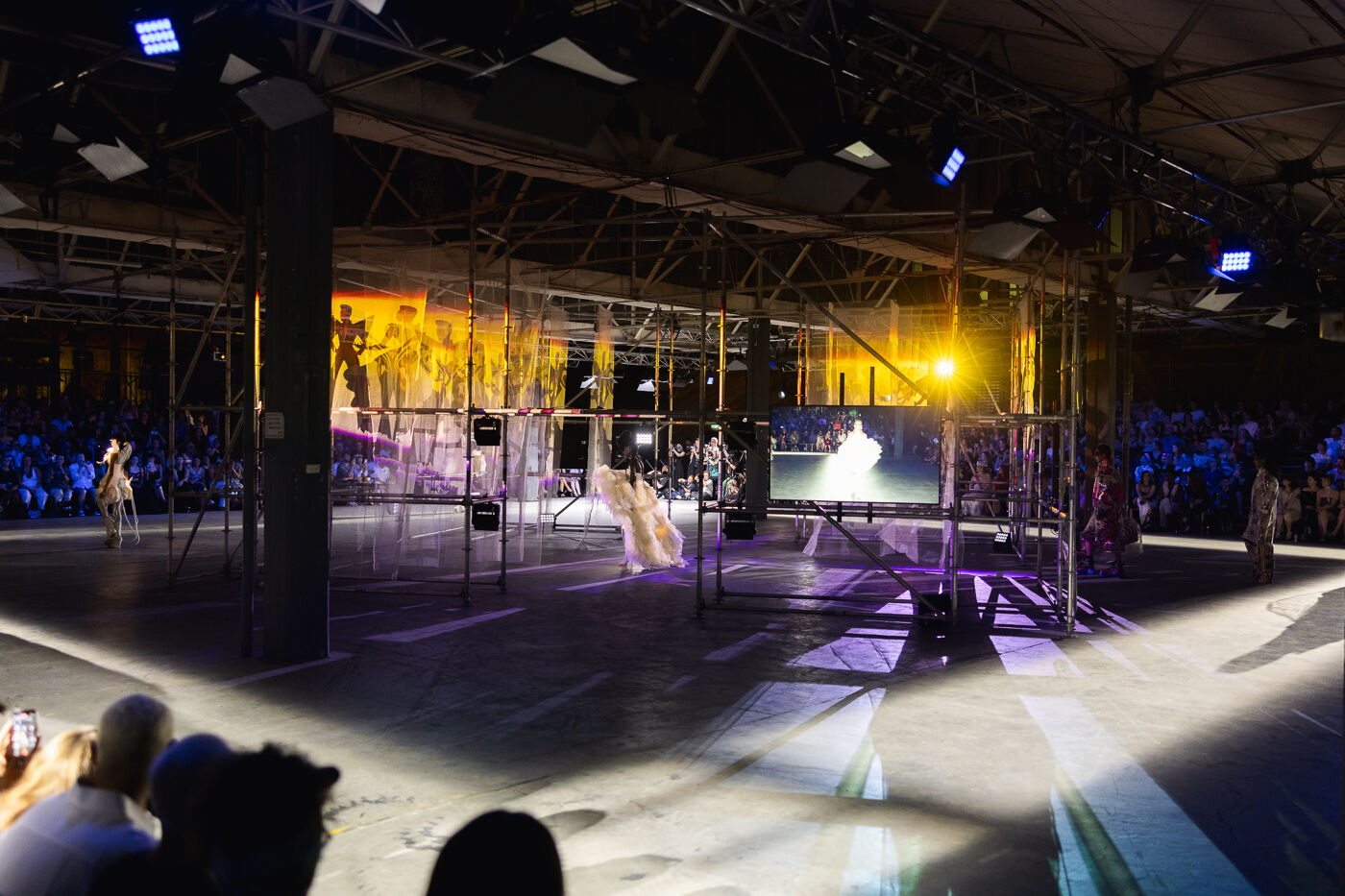 This image captures a fashion show with a modern, industrial aesthetic. Models walk down a runway surrounded by a metallic structure and screens displaying visuals, under dramatic lighting.