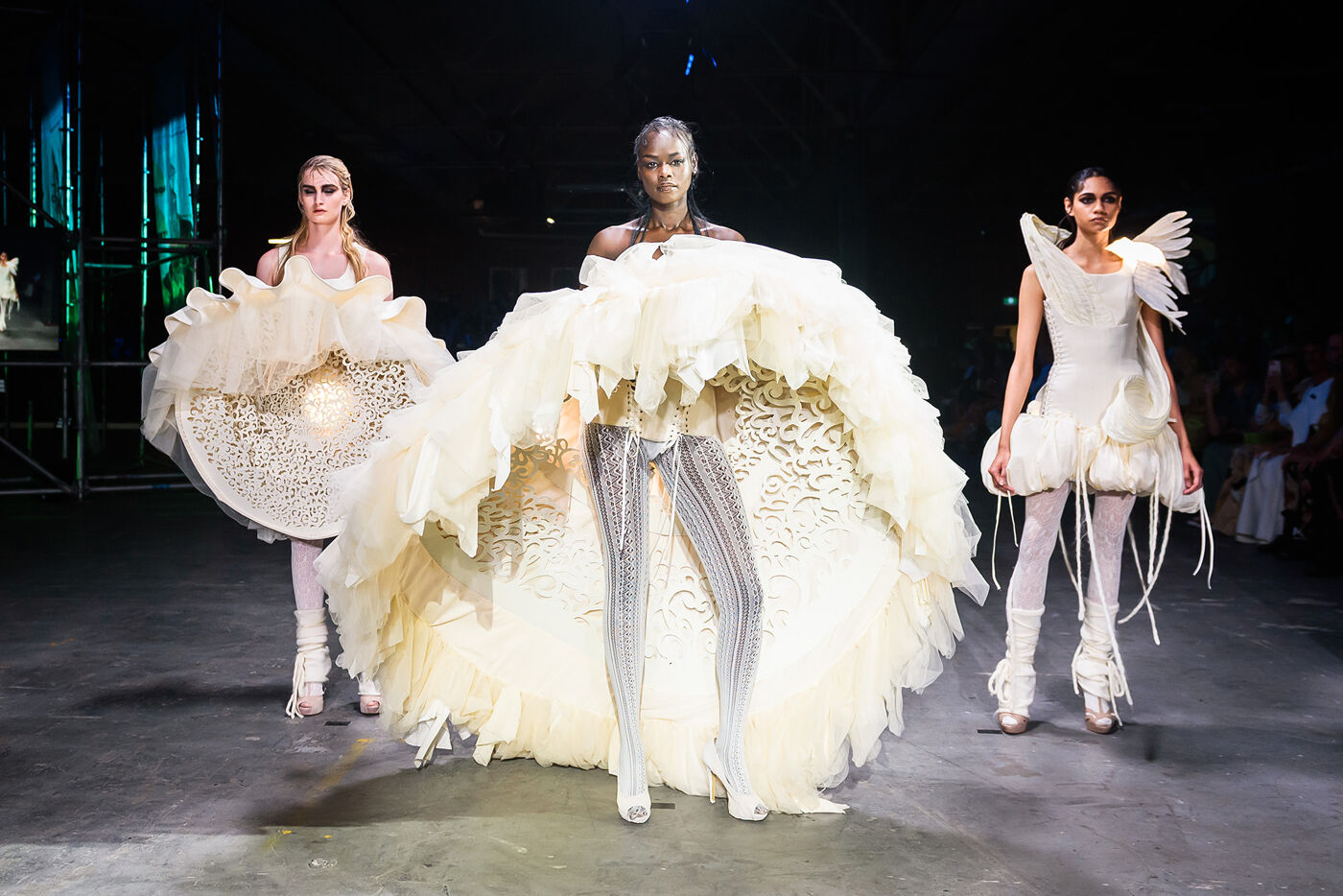 Three models are showcasing avant-garde, voluminous white dresses with intricate details on a dark runway, highlighting dramatic and artistic fashion design.