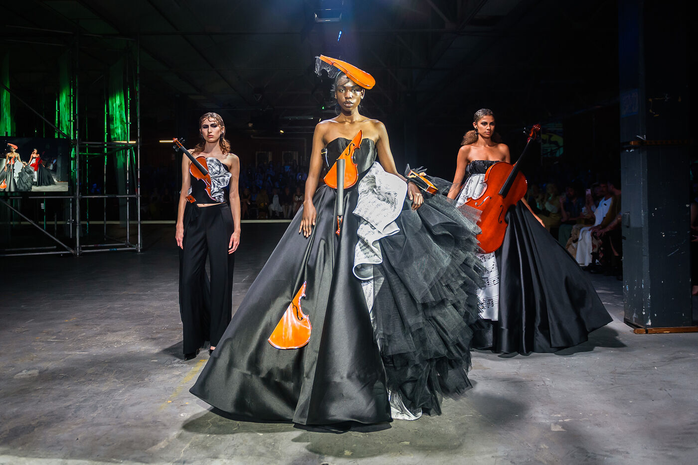 Three models walk down a runway showcasing avant-garde outfits inspired by musical instruments, featuring dramatic silhouettes and vibrant orange accents.