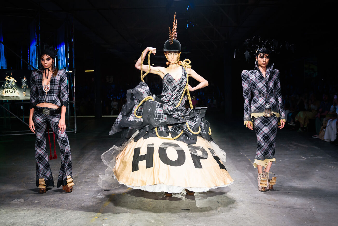 The image displays a fashion show with three models on a runway. The central model wears a voluminous gown with "HOPE" written on it, while the other two models are dressed in matching plaid outfits.