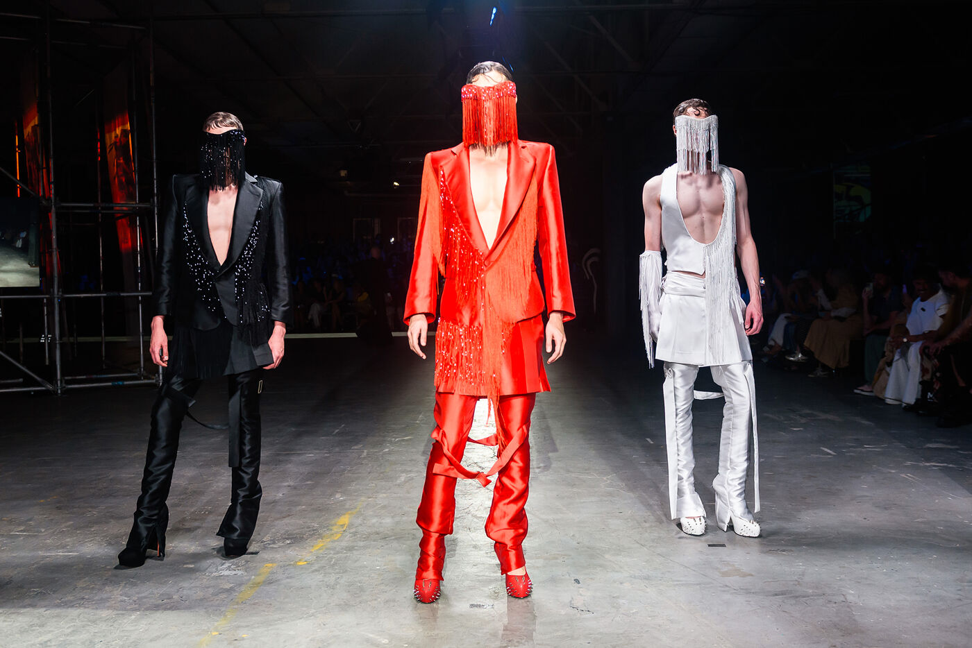 Three models walk the runway in avant-garde outfits. The outfits feature bold colors like red and black, with dramatic headwear and accessories, showcasing a futuristic and edgy fashion aesthetic.
