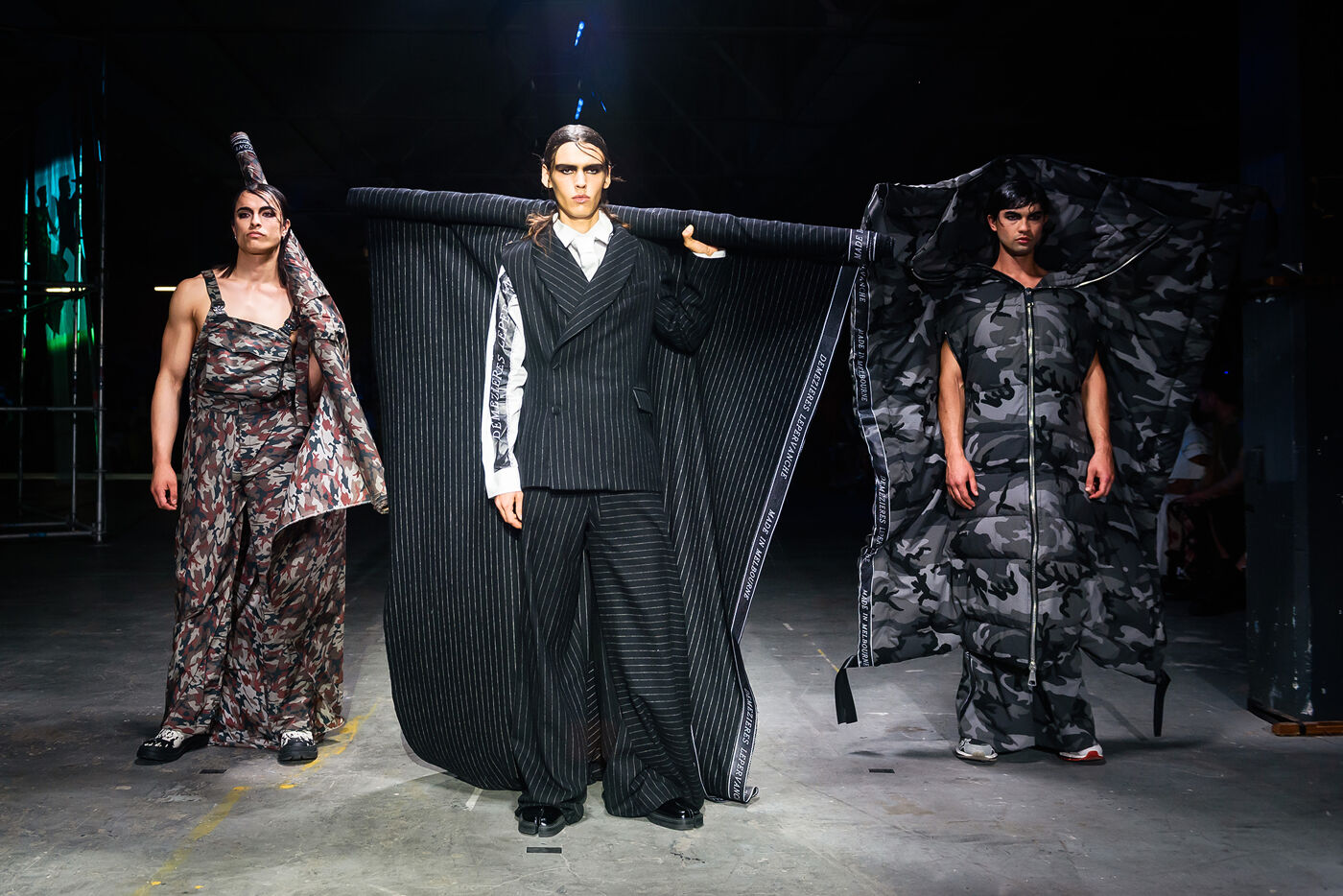 Three models walk the runway, showcasing avant-garde outfits. The central model wears a pinstripe suit with a dramatic cape, while the others are in patterned and camouflage ensembles.