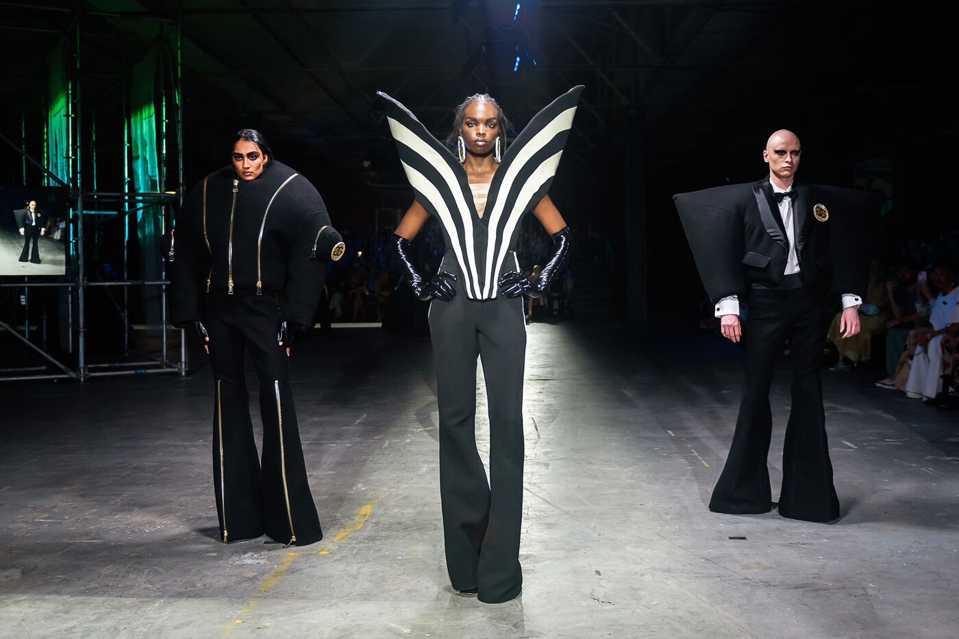 Three models walk down a runway in avant-garde black and white outfits. The central model wears a striking winged top, while the others are in dramatic, structured ensembles.