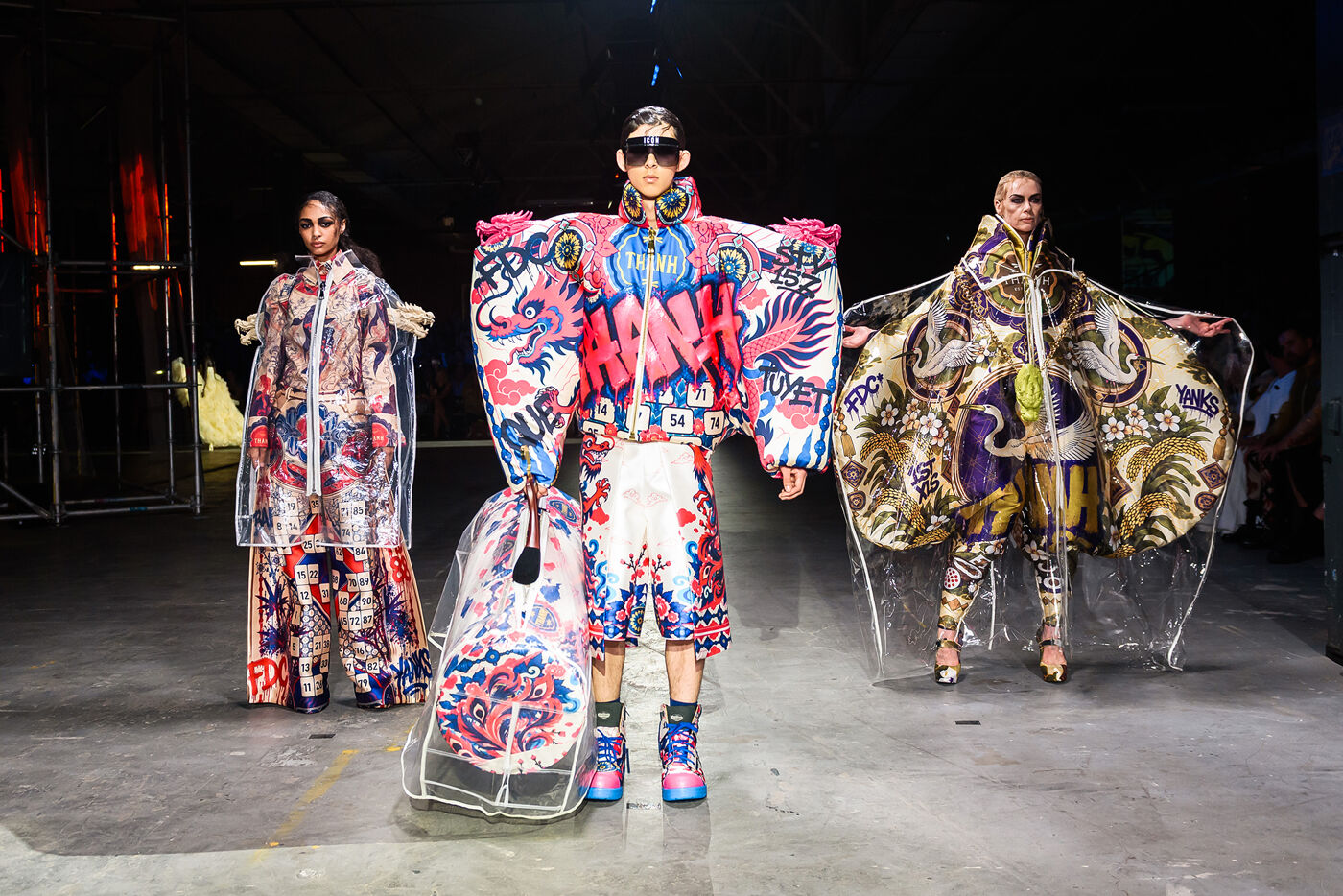 Three models walk down a runway showcasing avant-garde outfits featuring bold graffiti-inspired prints, oversized silhouettes, and transparent outerwear, presenting a vibrant and eclectic fashion statement.