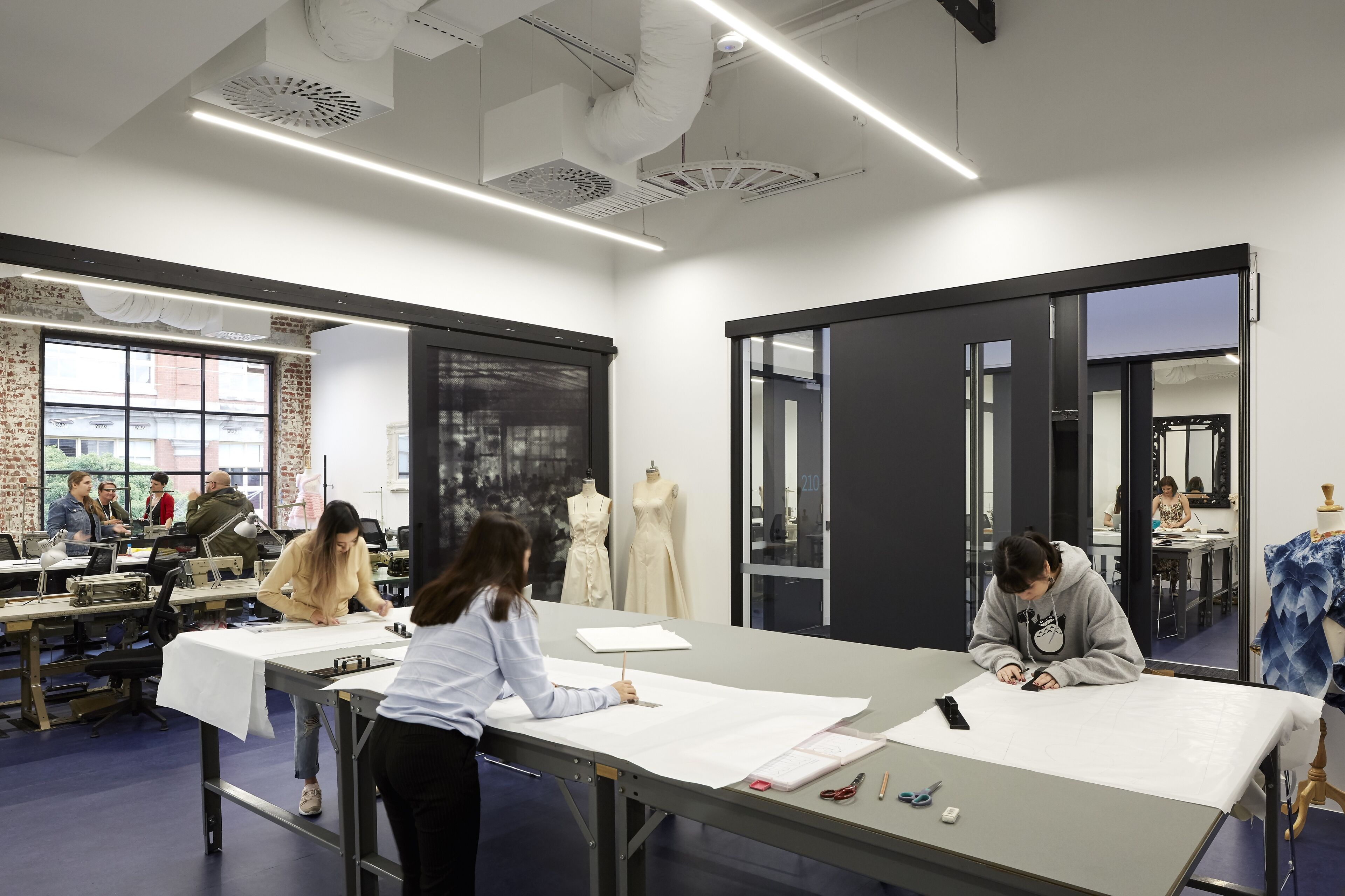 In the well-lit studio space, a sense of focused creativity fills the air. Students are engrossed in their tasks, drafting patterns and stitching fabric, surrounded by mannequins draped in half-completed designs. The exposed brick wall adds a touch of industrial charm, contrasting with the meticulous order of design tools and materials. It's a snapshot of dedication and the quiet buzz of potential.