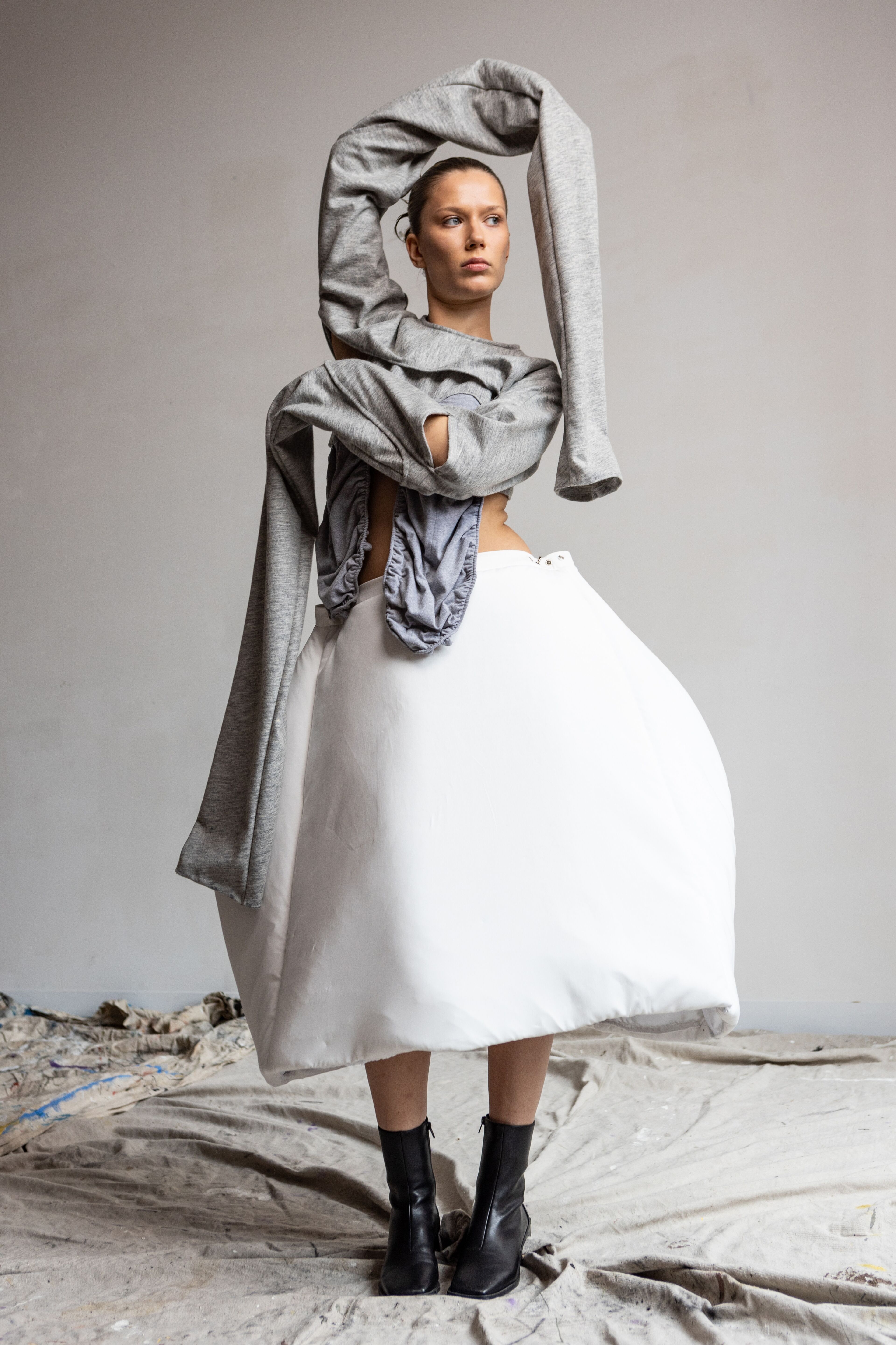 A bold fashion statement is captured in this image where a model stands in a striking pose, a voluminous white skirt ballooning around her, contrasted with a textured grey top that wraps around her arms.