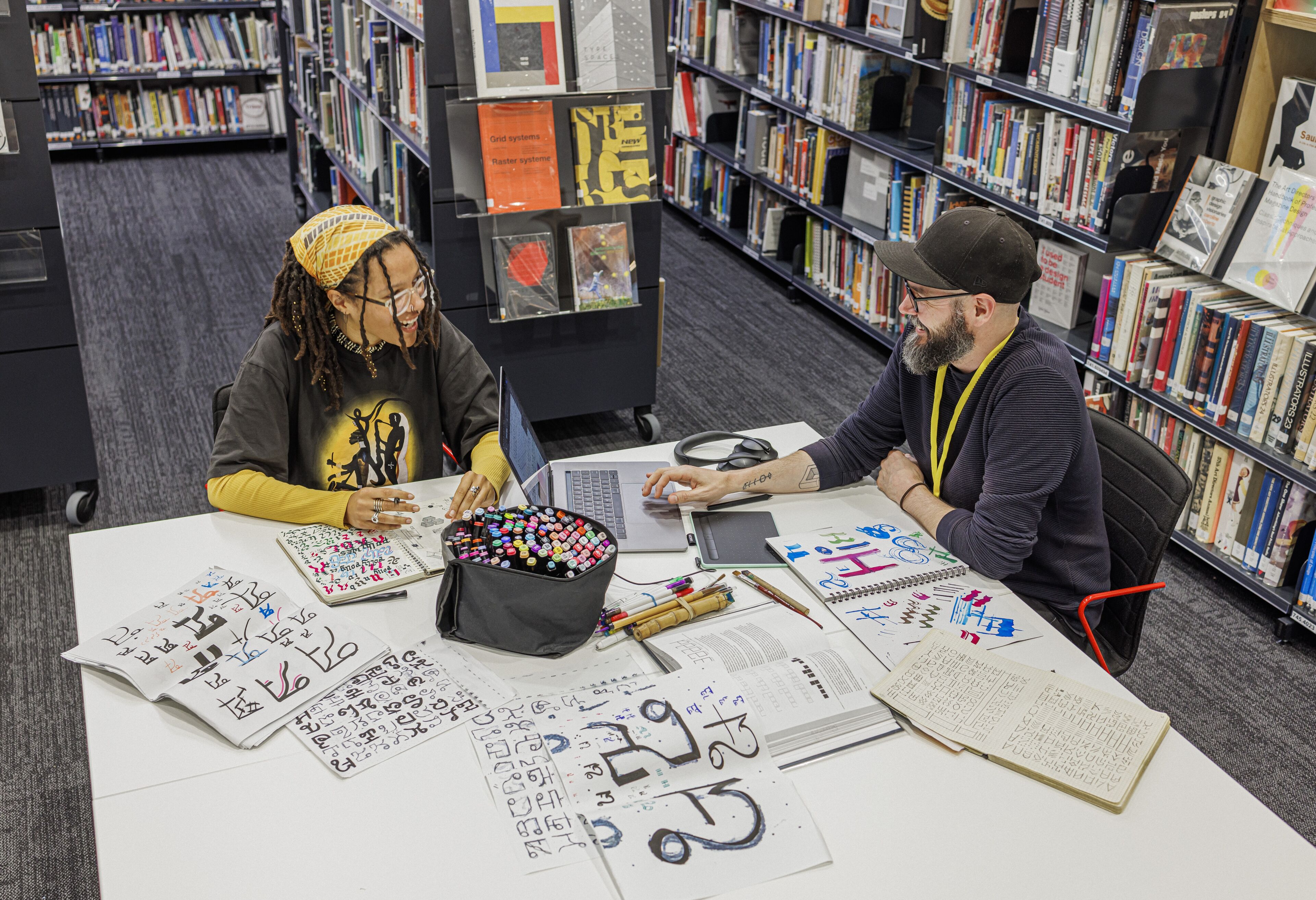 Two designers share a laugh over a table scattered with colorful markers and typography drafts in a well-stocked library.