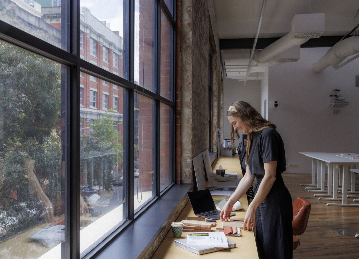 A designer arranges materials on a desk bathed in natural light, with a vibrant cityscape outside.