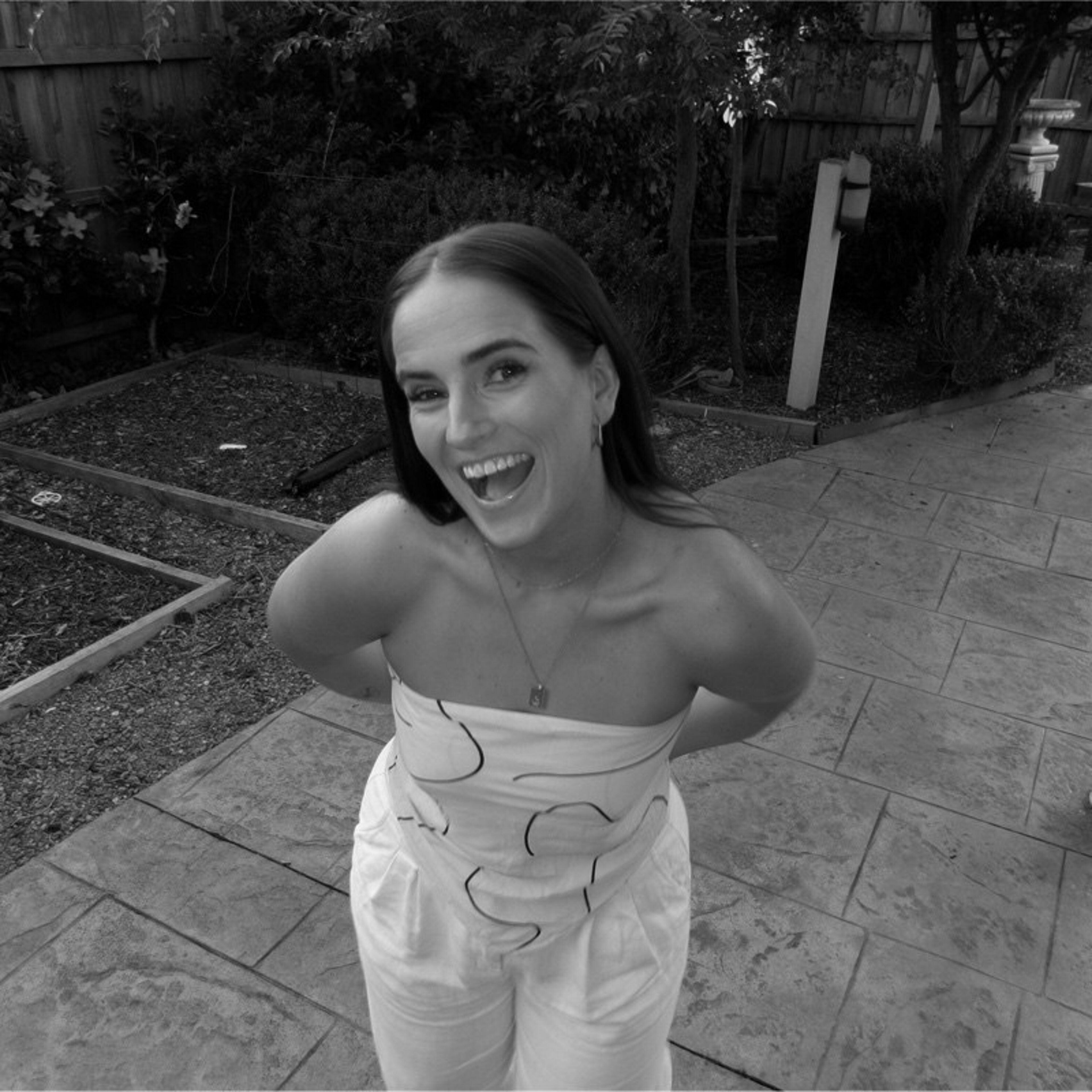 Monochrome image of a joyful woman in a strapless dress smiling broadly, standing in a residential garden.