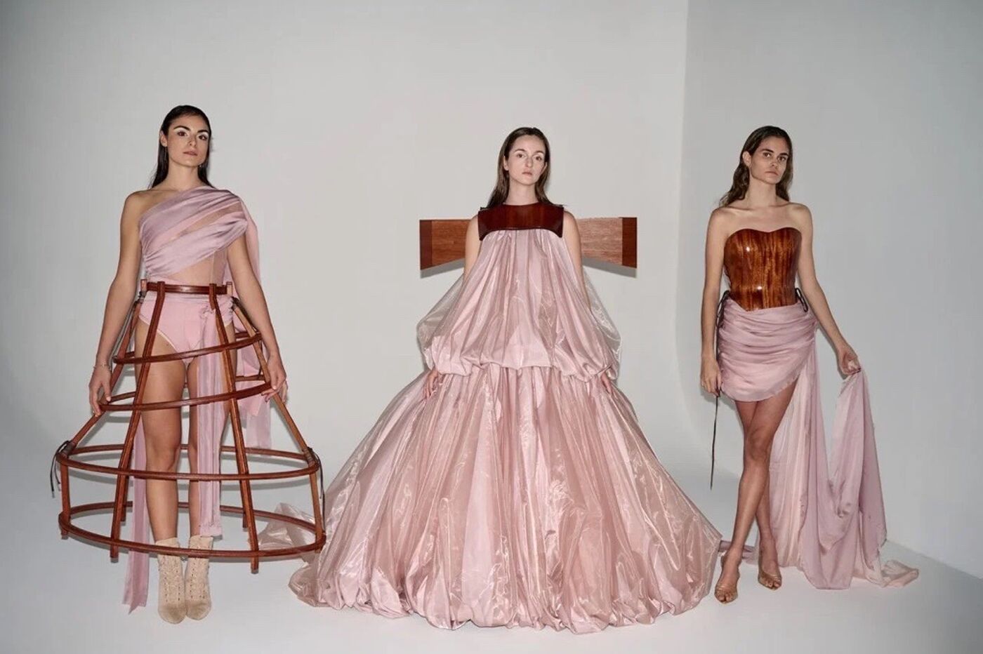 The image showcases three models in avant-garde fashion. Each model wears a unique, flowing gown with distinct structural elements and a soft color palette, creating a visually striking composition.