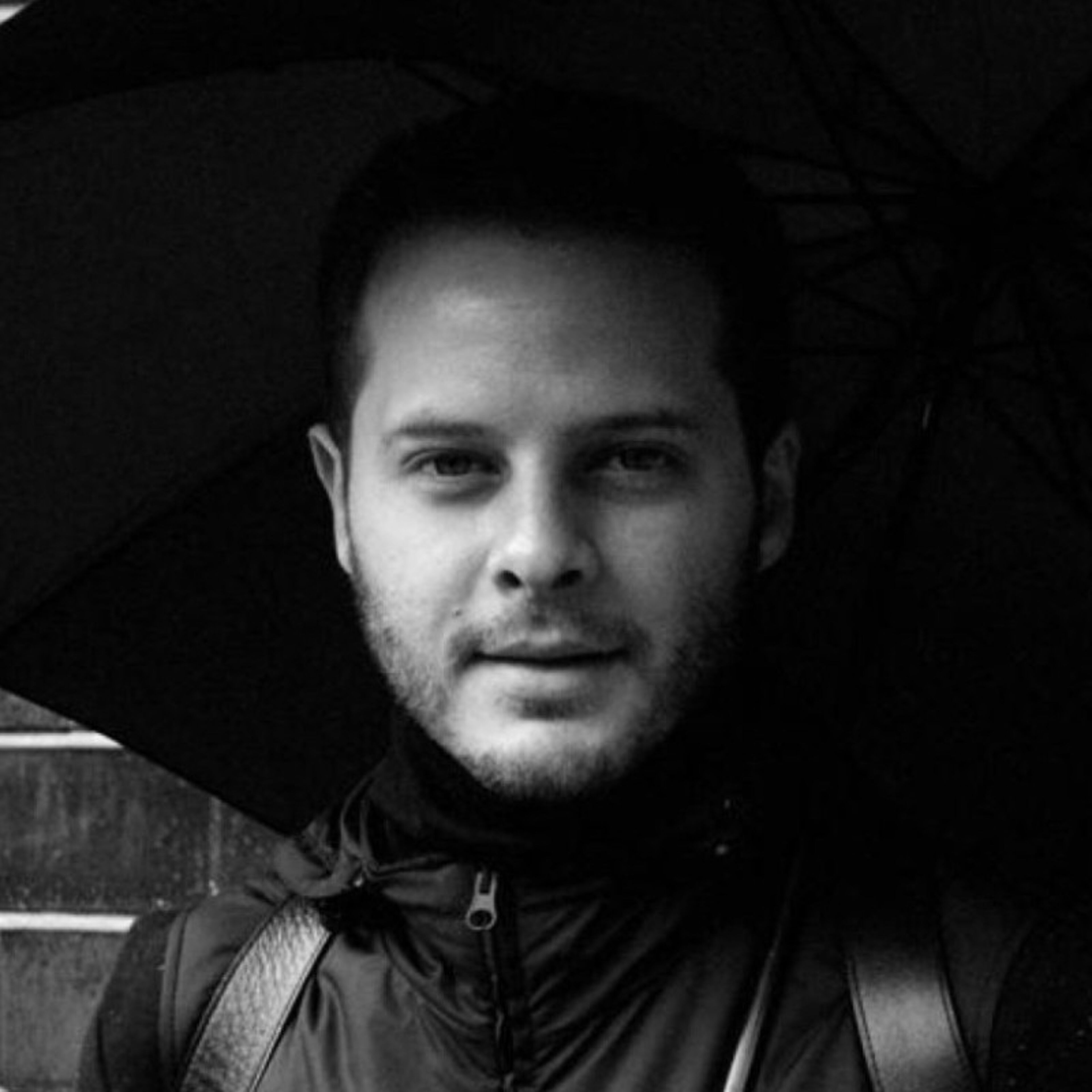 A monochrome portrait of a man holding an umbrella, gazing directly at the camera with a subtle smile.