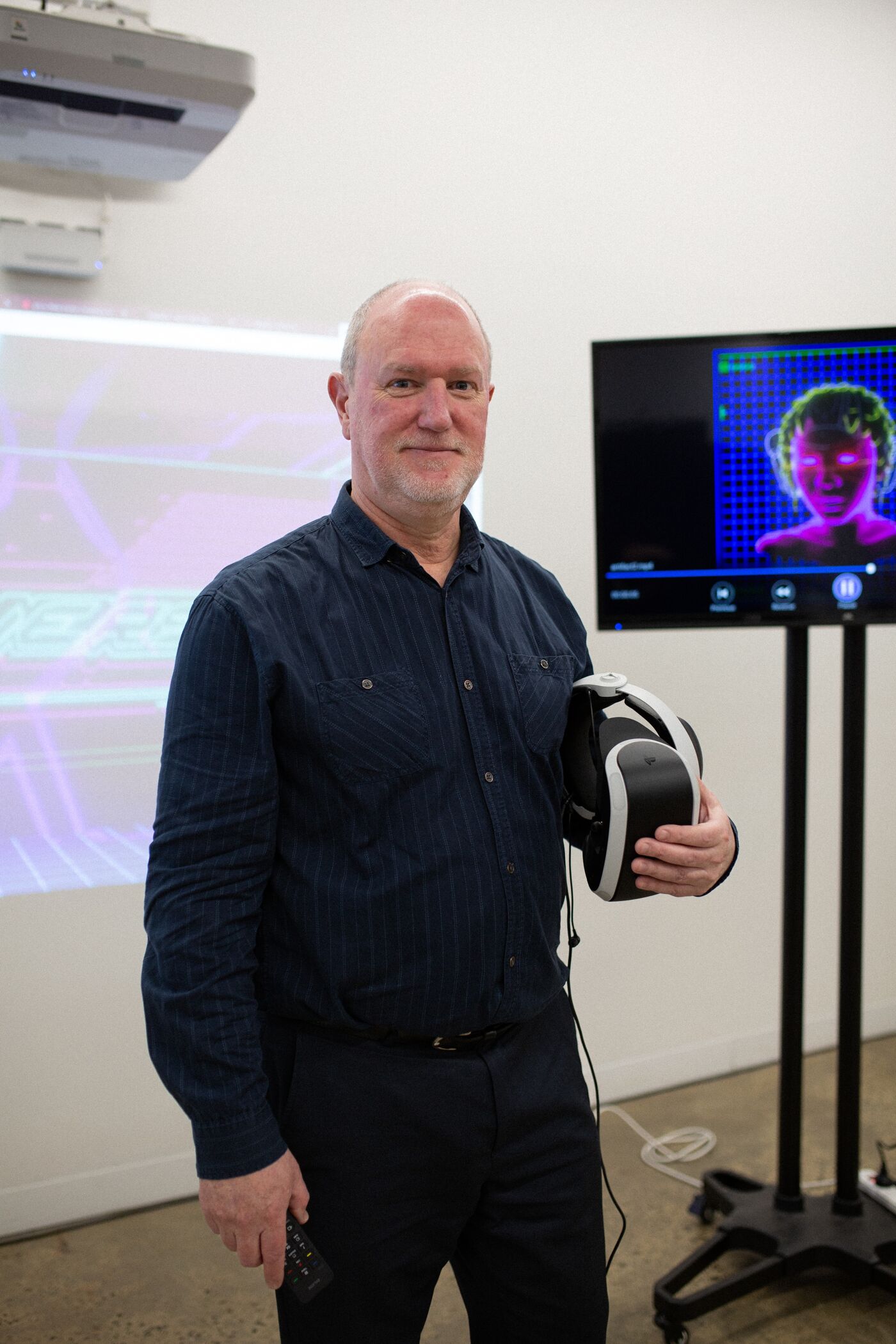 A man stands with a VR headset, remote in hand, in front of a projection screen at a technology presentation, illustrating the hands-on aspect of immersive experiences.