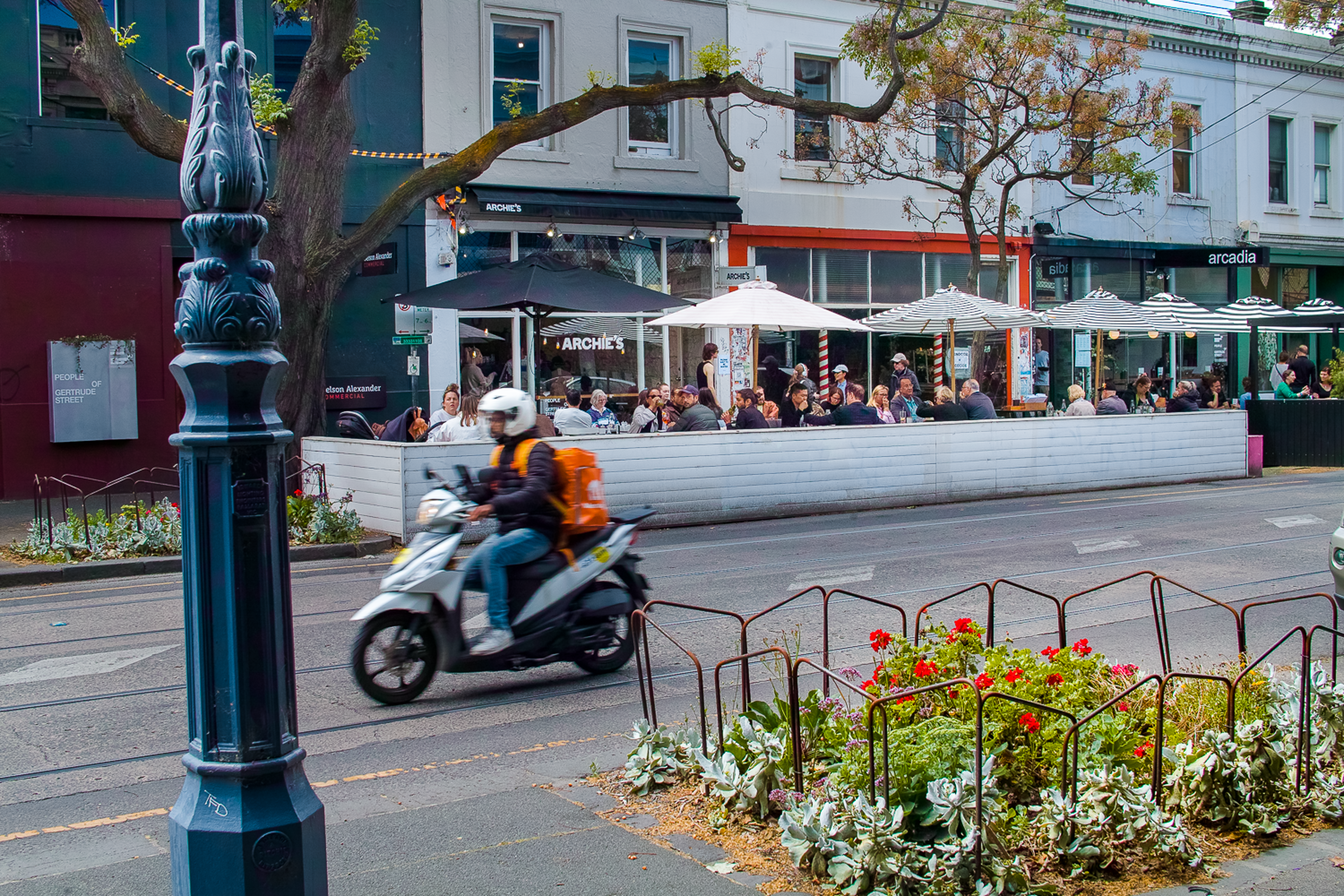 A bustling street scene featuring an outdoor cafe with patrons under striped umbrellas, framed by a decorative street lamp and vibrant flower beds, as a motorcyclist zooms by.