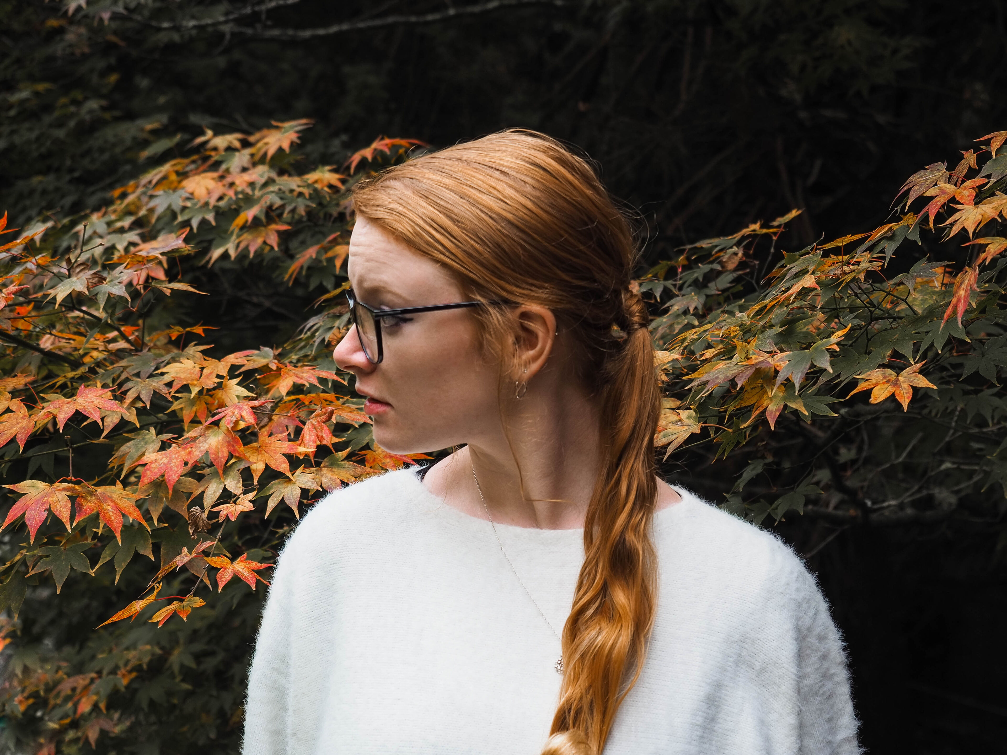 A red-haired woman in profile with glasses, wearing a white sweater, stands before a backdrop of autumn leaves.