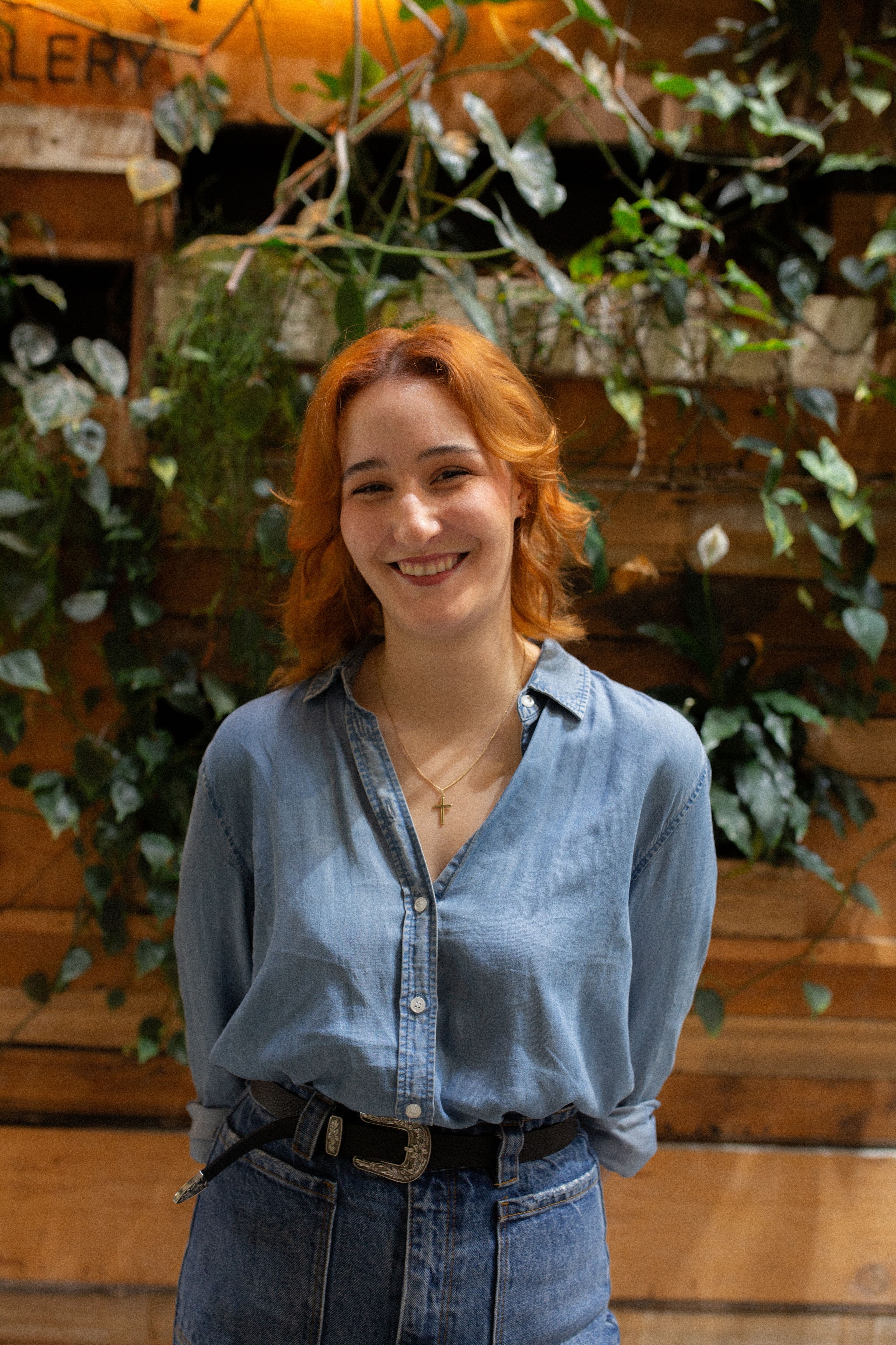 A cheerful young woman with shoulder-length red hair, wearing a blue denim shirt and high-waisted jeans, stands in front of a leafy backdrop.