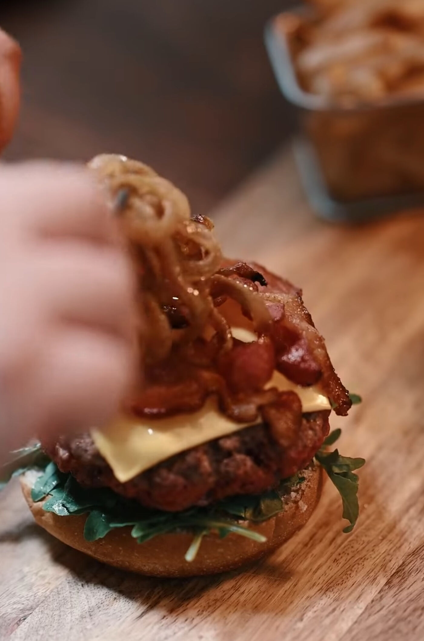 A close-up of a gourmet burger being assembled on a wooden board. The layers include fresh arugula, a juicy beef patty, melted cheese, crispy bacon, and caramelized onions being added by hand. A container of fried onions is visible in the background.