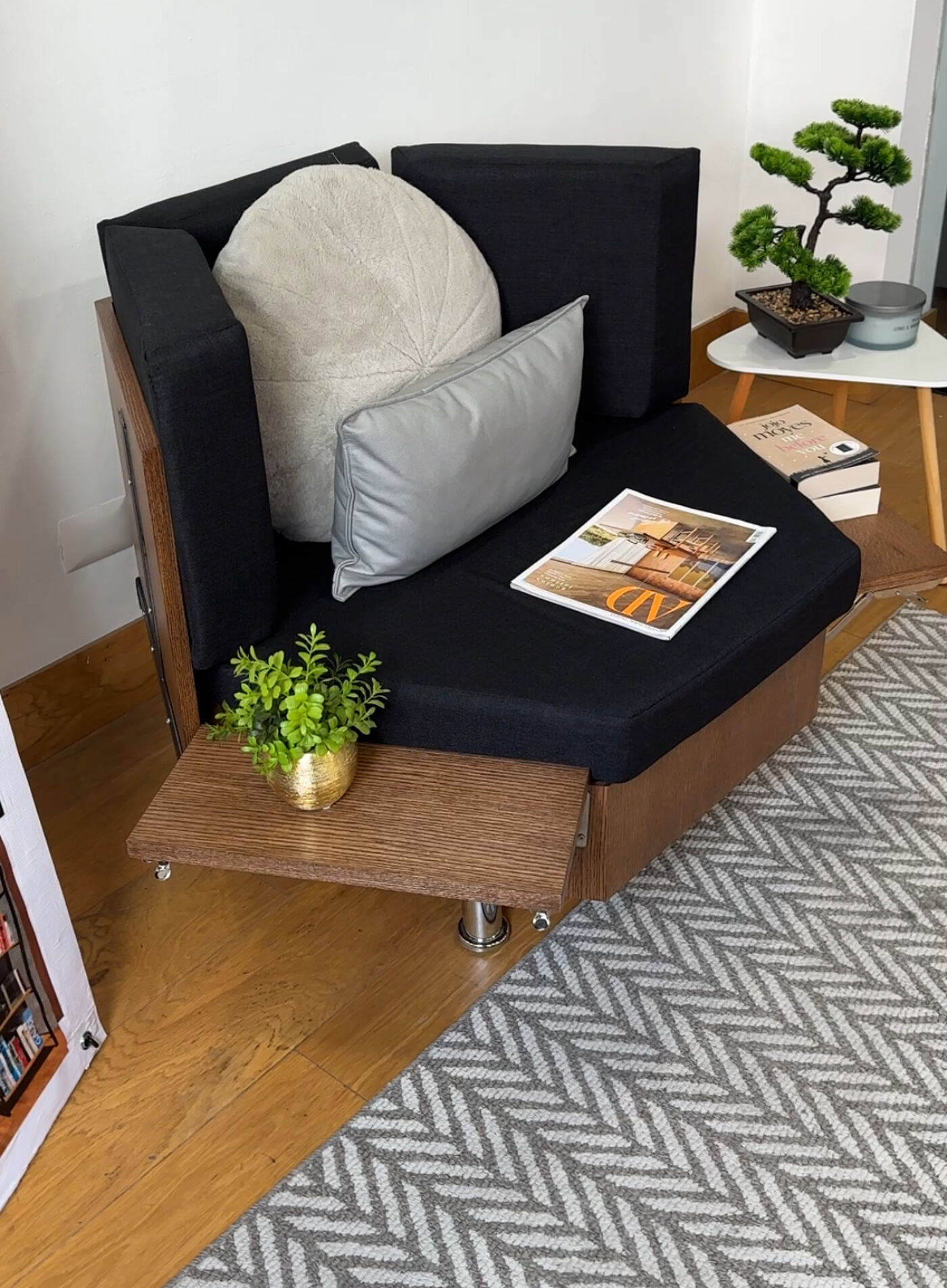 A black upholstered lounge chair with a cozy, minimalist design. It features a built-in wooden side table holding a small potted plant. The setting includes decorative cushions, a bonsai tree on a side table, and a patterned rug on a wooden floor.