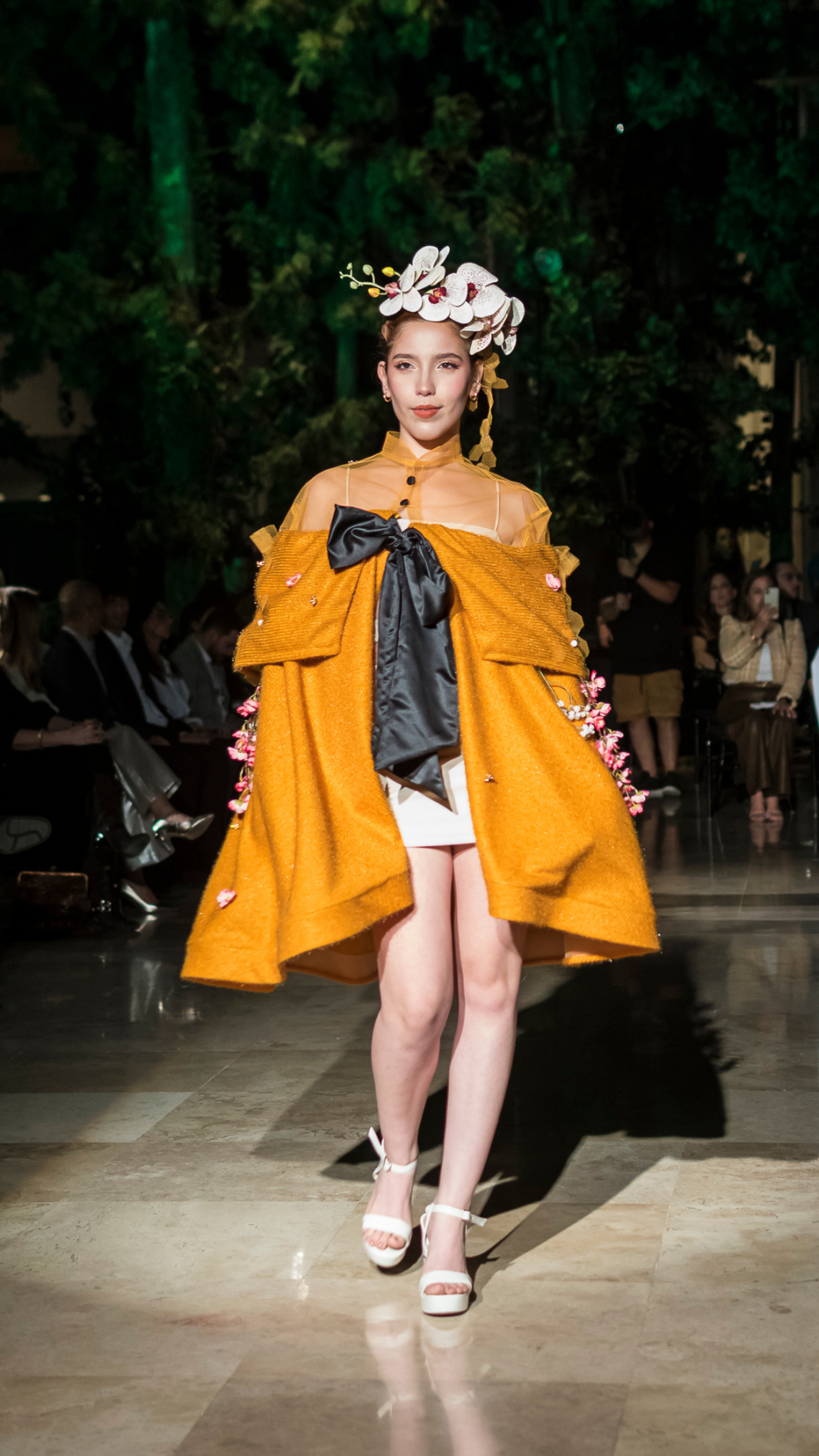 A young woman in a mustard-colored coat and white sandals walks down a runway, wearing a white orchid headpiece.