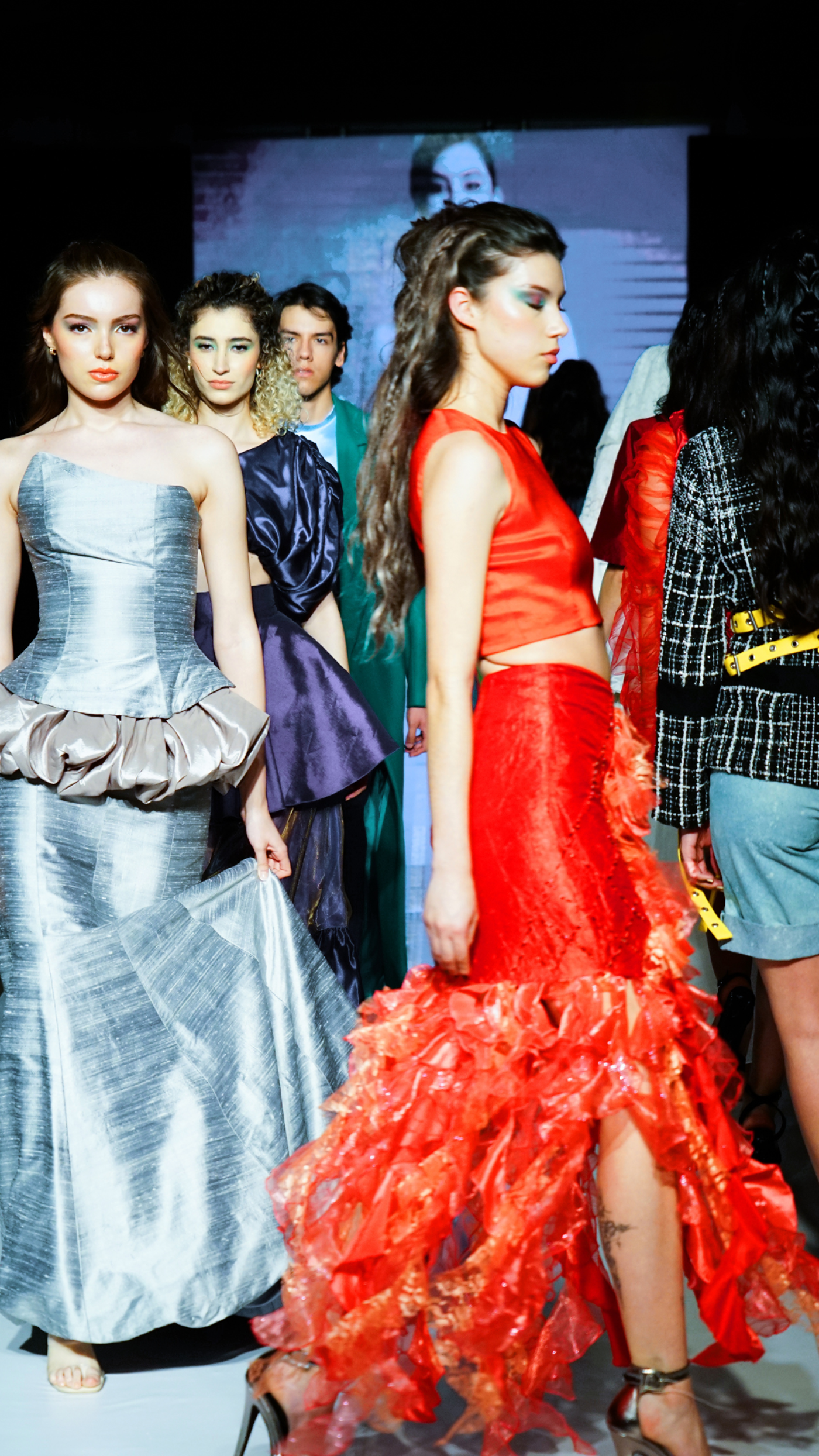 A group of models walk a runway showcasing various dresses, including a silver gown, a purple dress, and a vibrant red ruffled skirt.