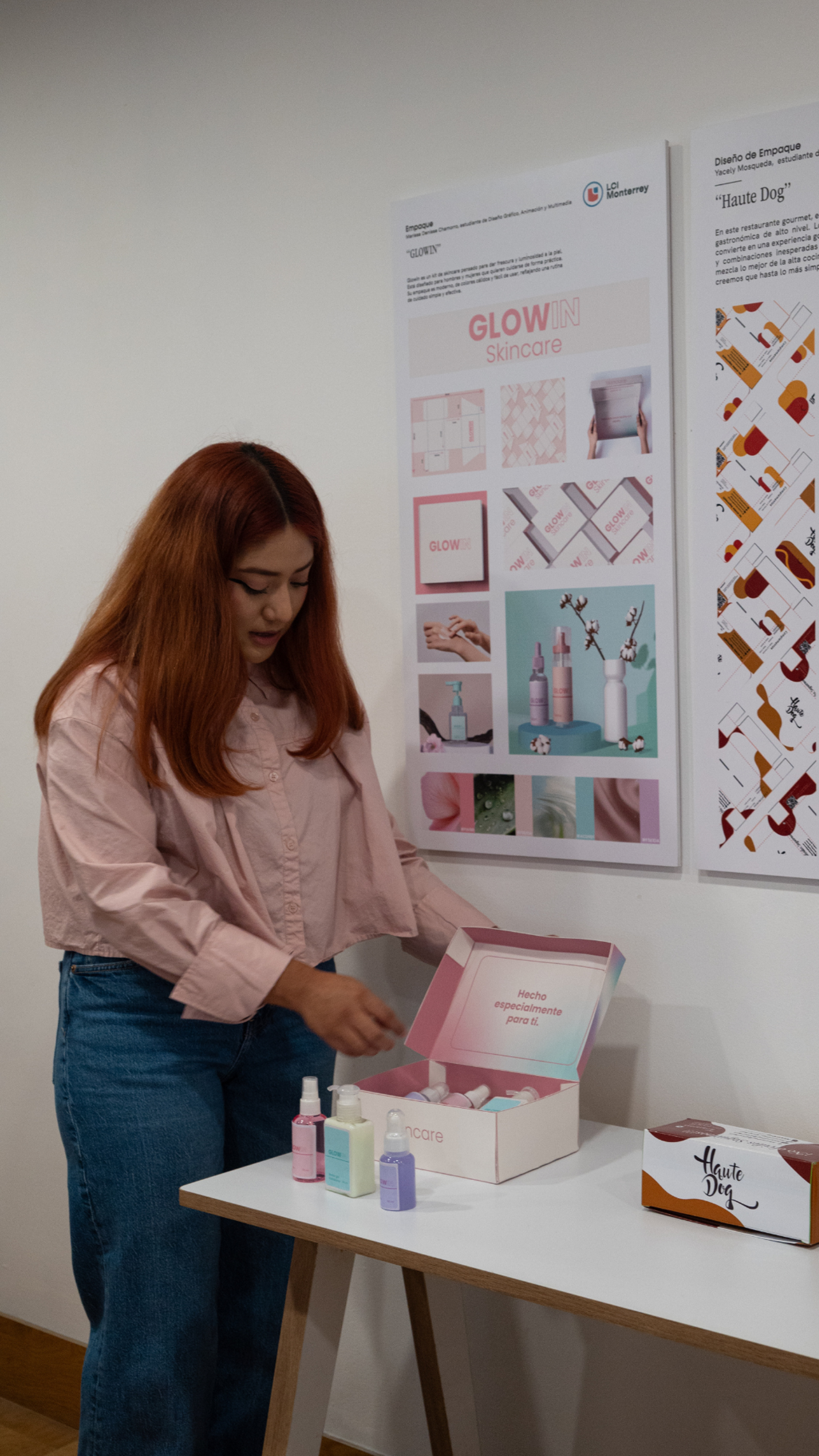 A young woman with long reddish-brown hair is standing at a small white table, holding open a box of skincare products, with design boards showcasing the product line in the background.