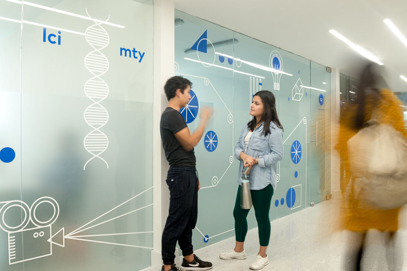 Two students converse by a decorative glass wall filled with scientific and mathematical icons.