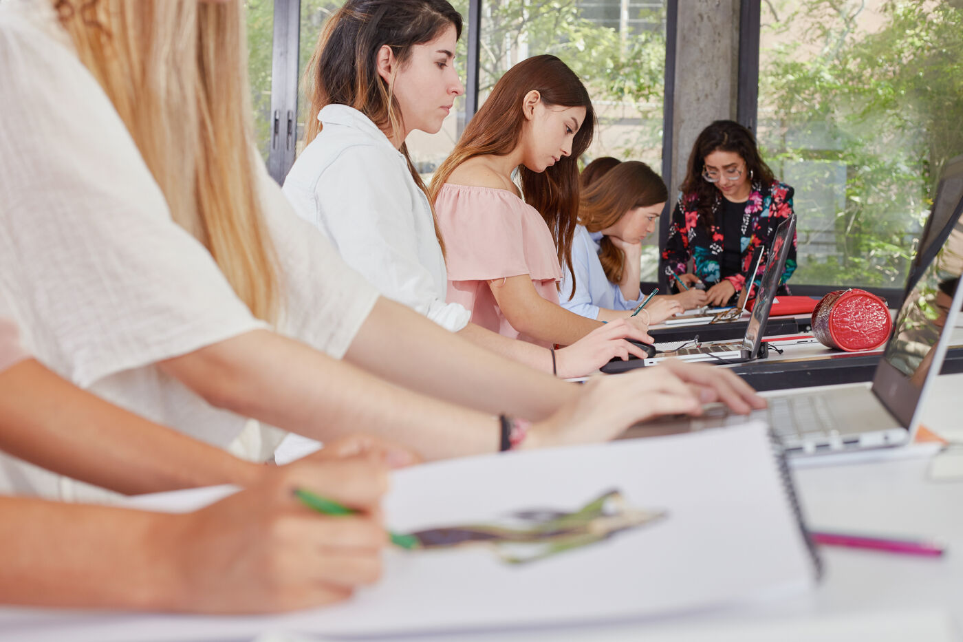 Focused students work together, with notebooks and laptops, in a bright classroom filled with natural light.