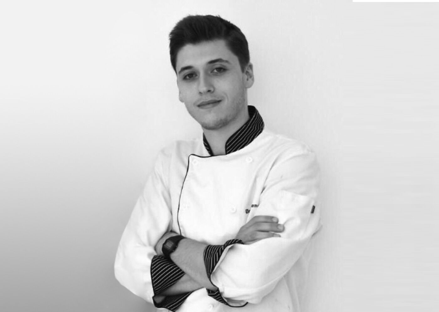 A monochromatic portrait of a young male chef in a white double-breasted jacket with folded arms, exuding confidence.