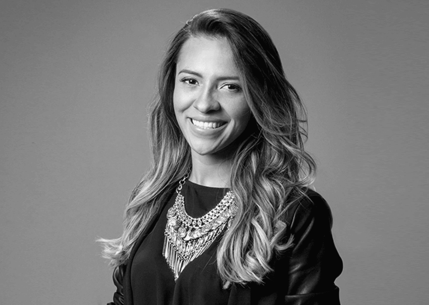 A confident businesswoman in a black blazer and ornate necklace smiles warmly, exuding professionalism and approachability in a corporate setting.