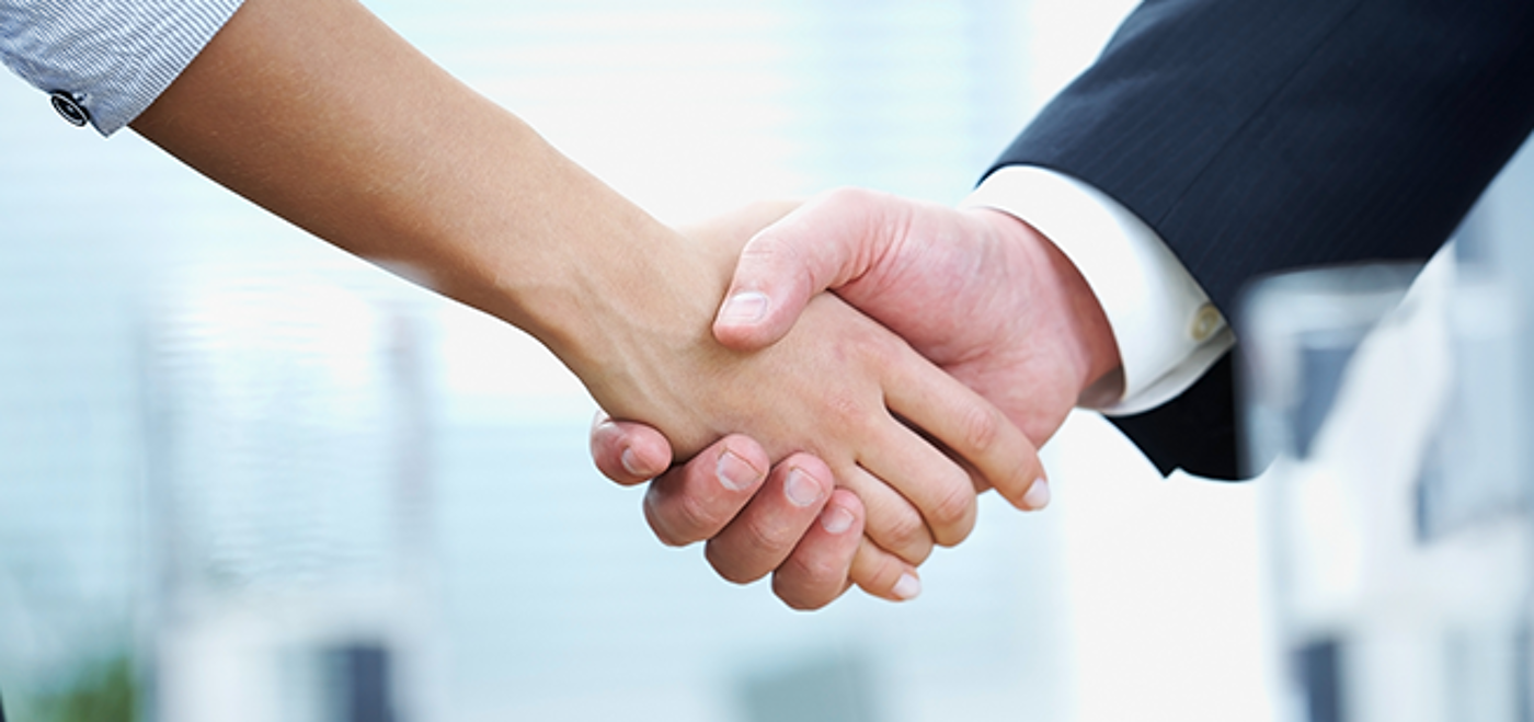 Two professionals, one wearing a light gray shirt and the other in a dark suit, engaging in a handshake in front of a blurred office background, symbolizing a successful agreement or partnership.