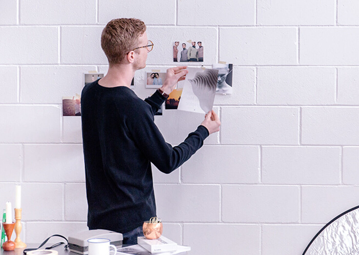 A young man in a black sweater arranges photographs on a mood board against a white brick wall in a creative workspace setting.
