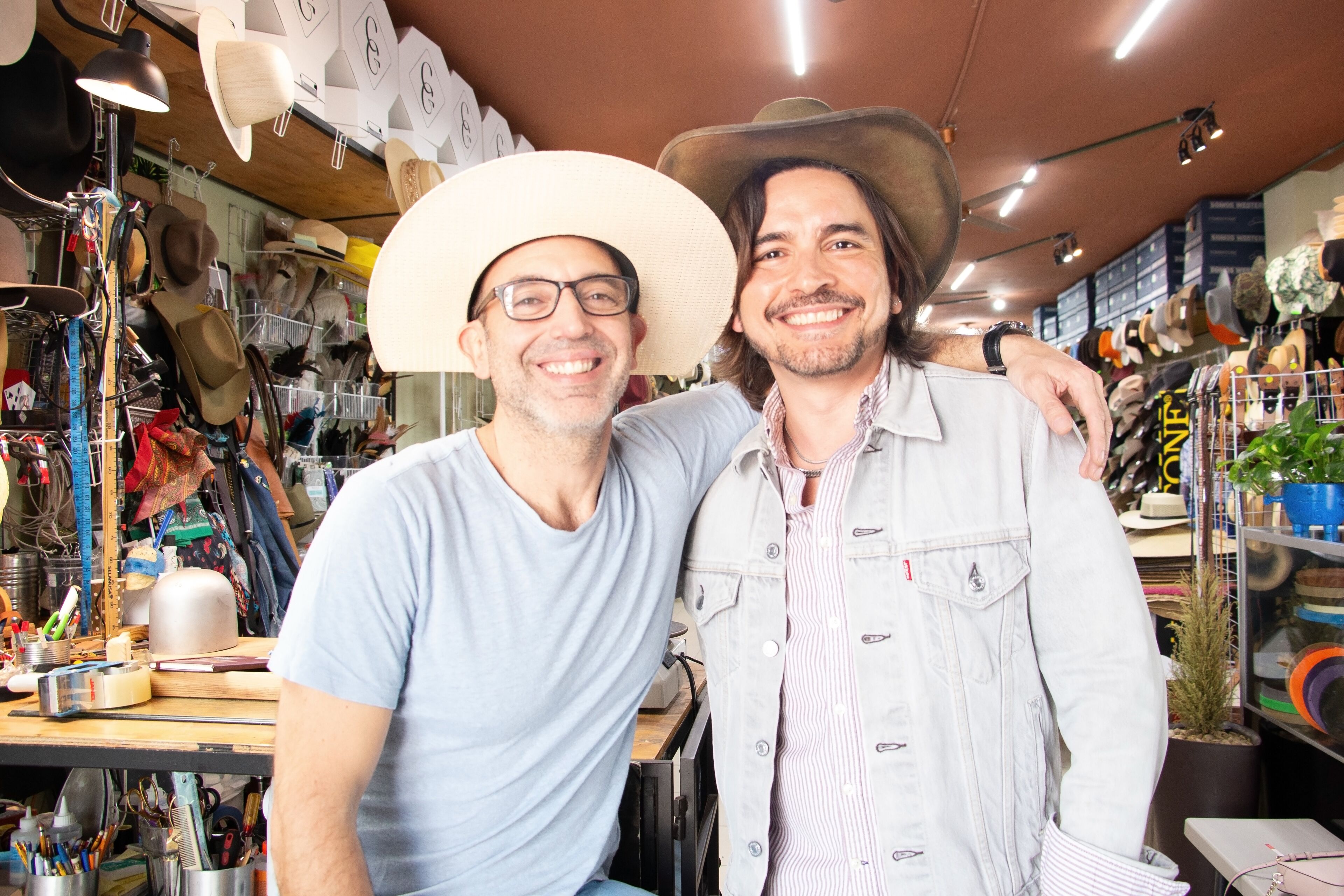Dos hombres sonrientes en una tienda de sombreros con varios estilos. Uno lleva un fedora blanco y el otro un sombrero de vaquero, mostrando un ambiente de negocio alegre.
